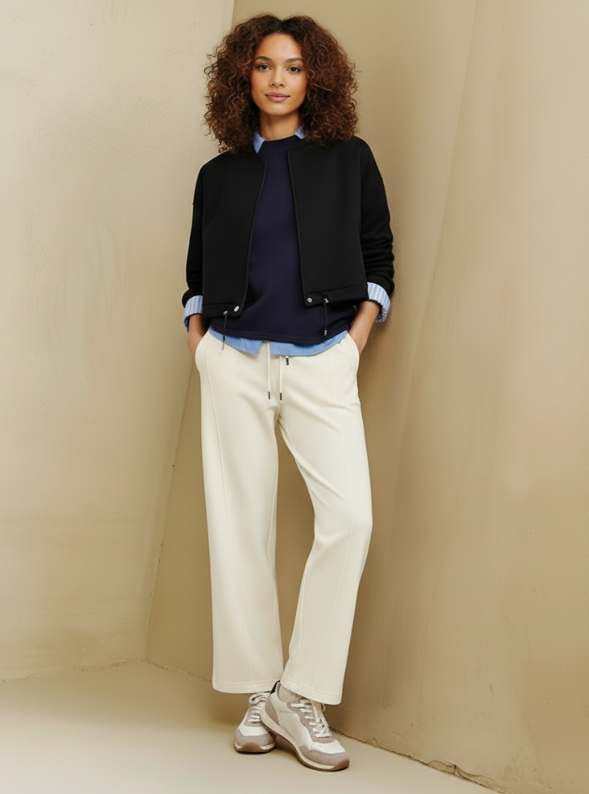 Woman wearing a black jacket, blue shirt, white pants, and sneakers against a beige wall.