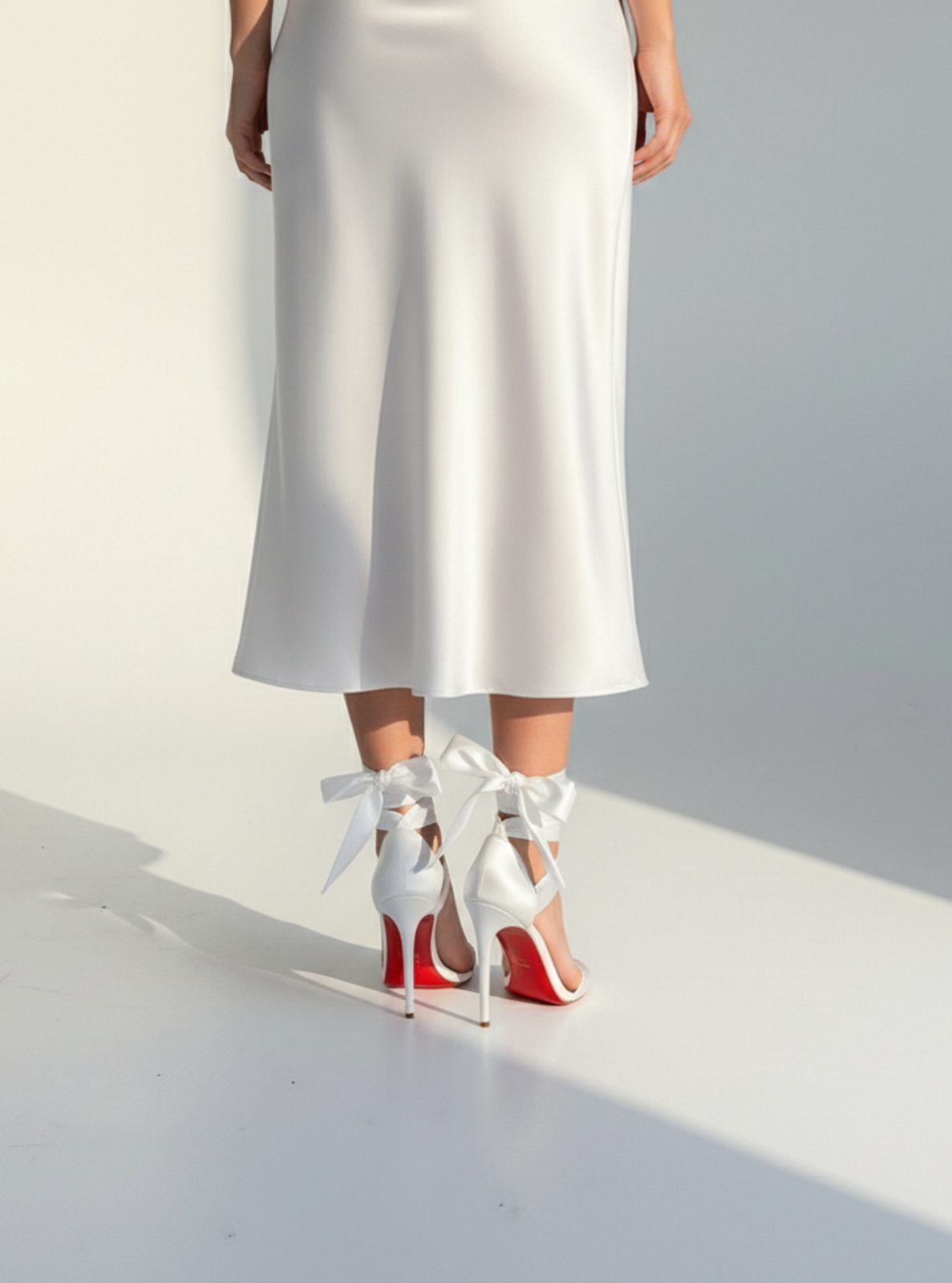 White high-heeled shoes with red soles and white bows worn by a person in a white dress on a light gray background.