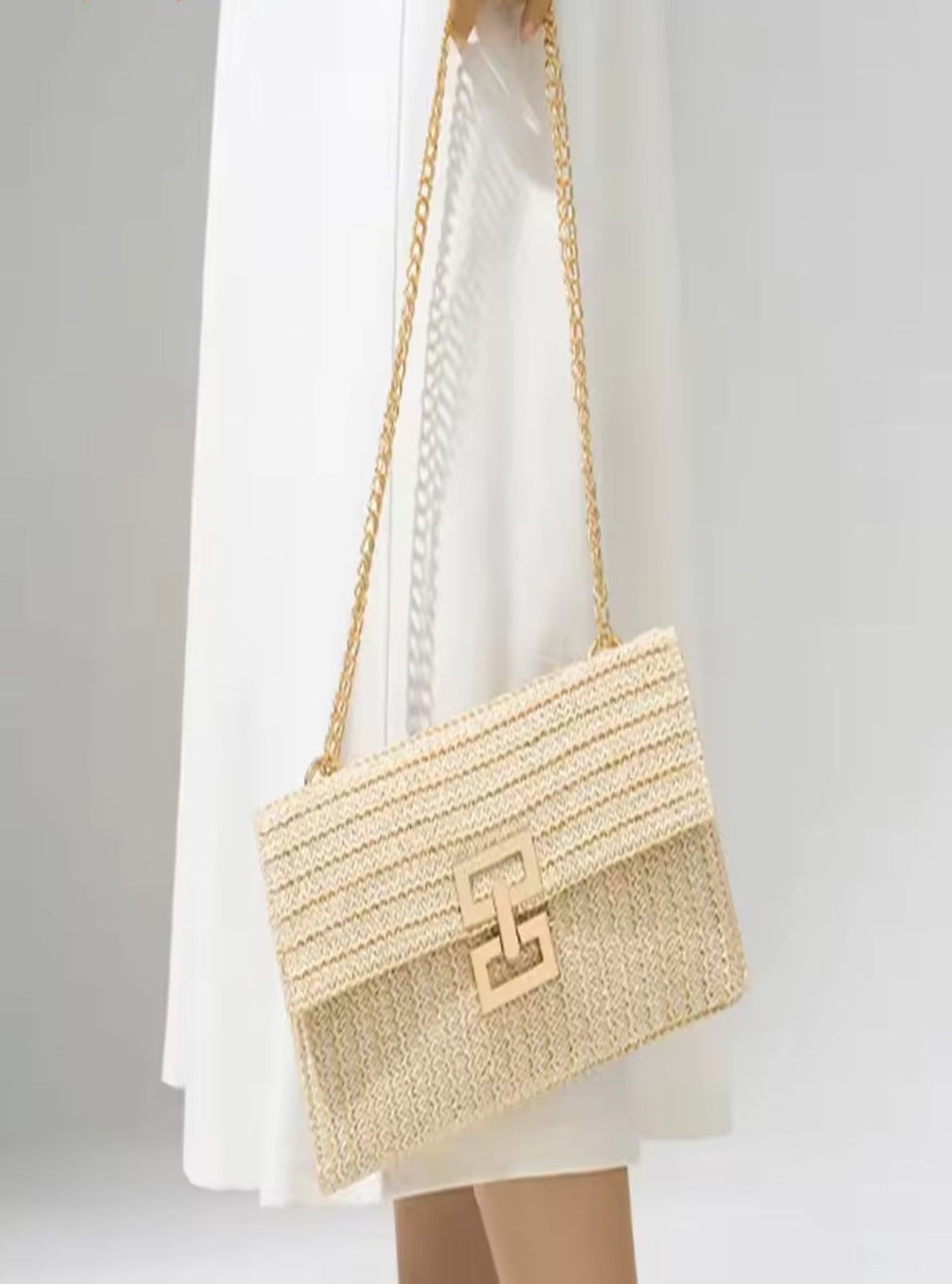 Beige handbag with a gold chain strap held by a person wearing a white dress.