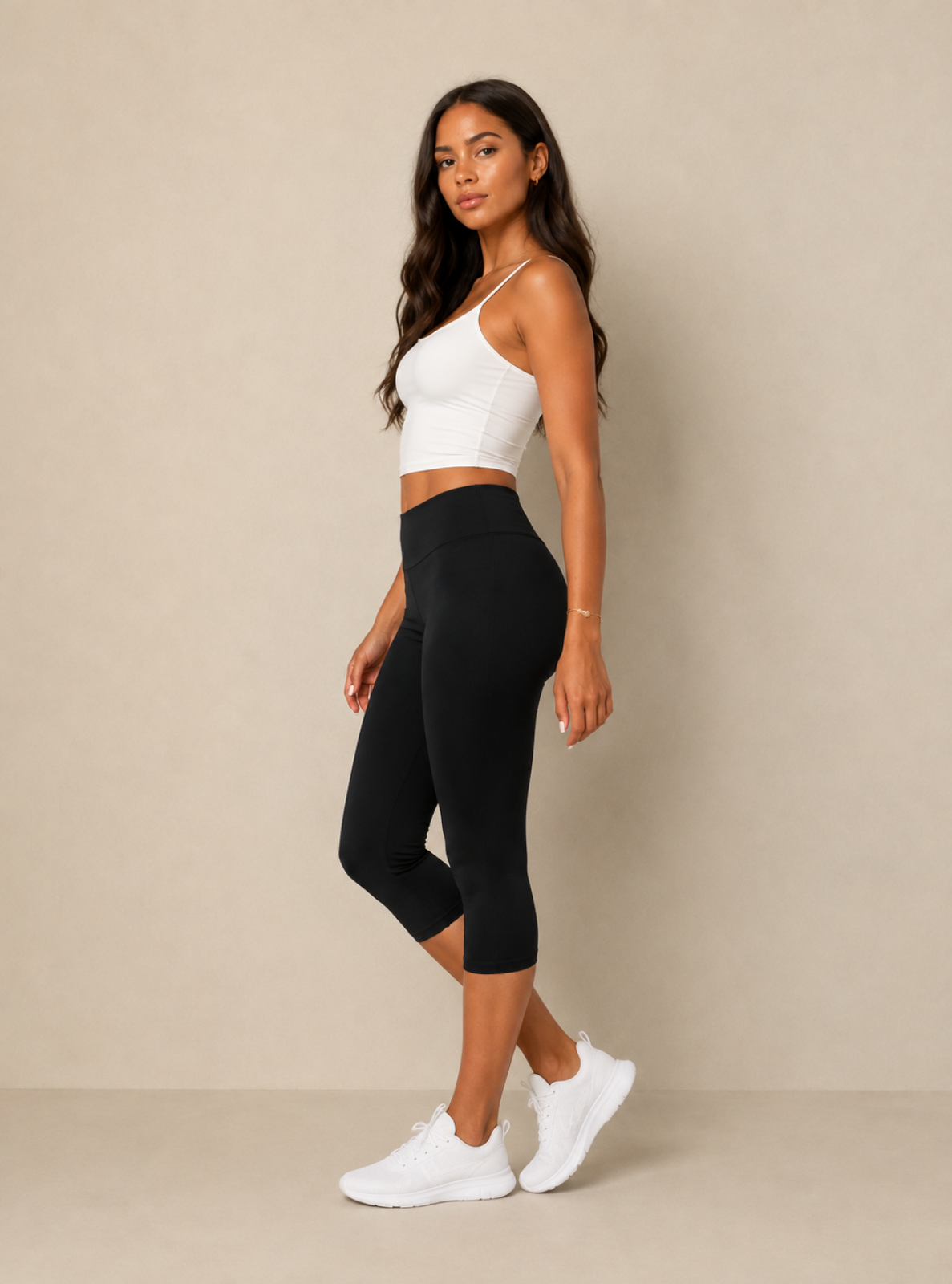 A woman modeling black high-waisted capri leggings, paired with a white ribbed crop top and white sneakers, standing against a neutral beige studio backdrop.