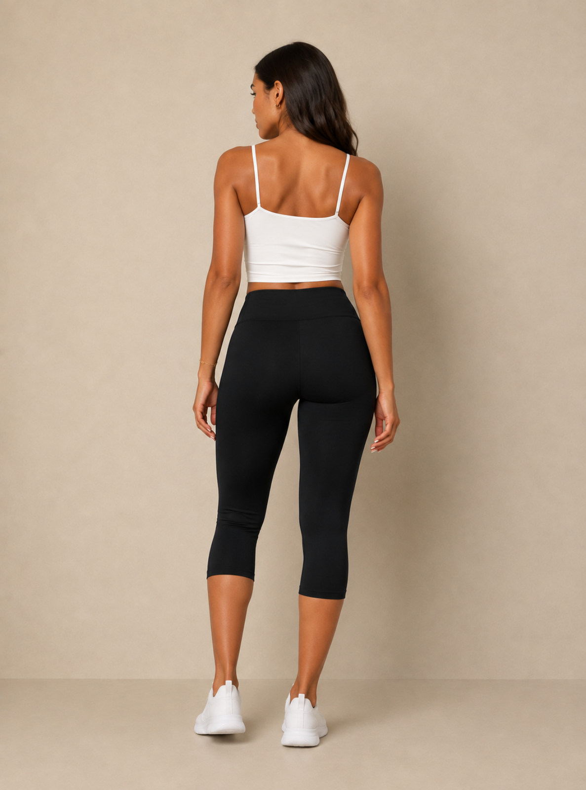 A woman is shown from the back wearing black high-waisted capri leggings, a white crop top, and white sneakers against a neutral beige studio backdrop.