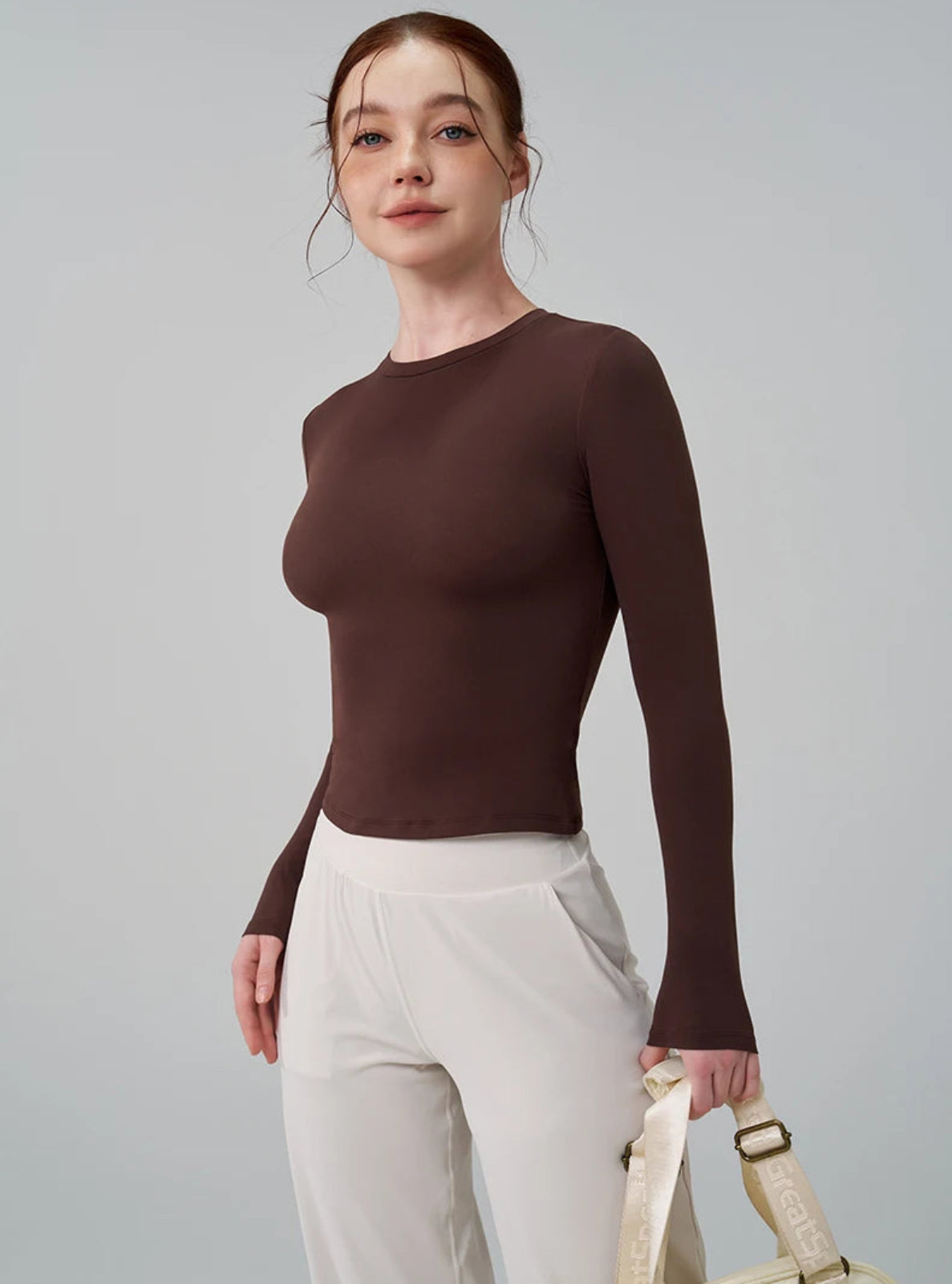 Woman wearing a brown long-sleeve top and white pants, holding a beige handbag against a gray background