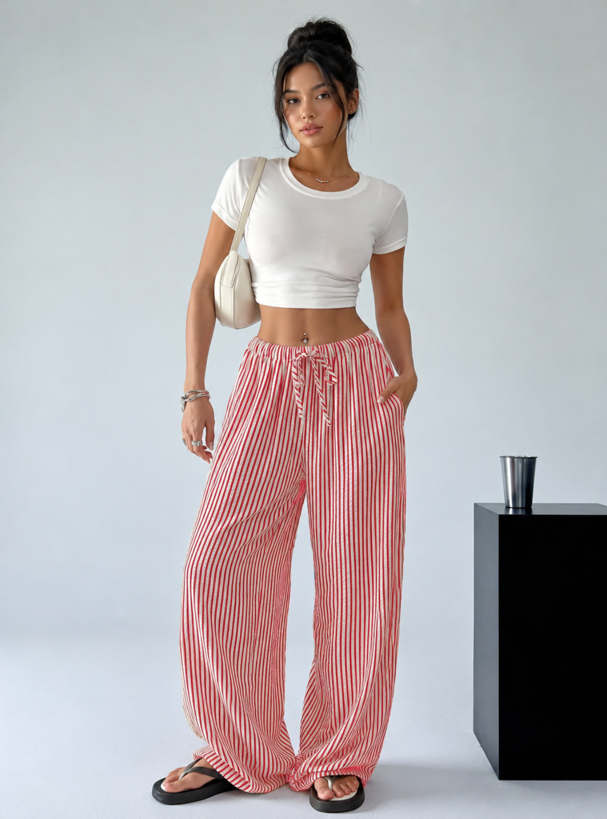 A woman modeling a white cropped short-sleeve t-shirt and pink-and-white vertically striped wide-leg drawstring pants, standing in a studio setting next to a black pedestal.