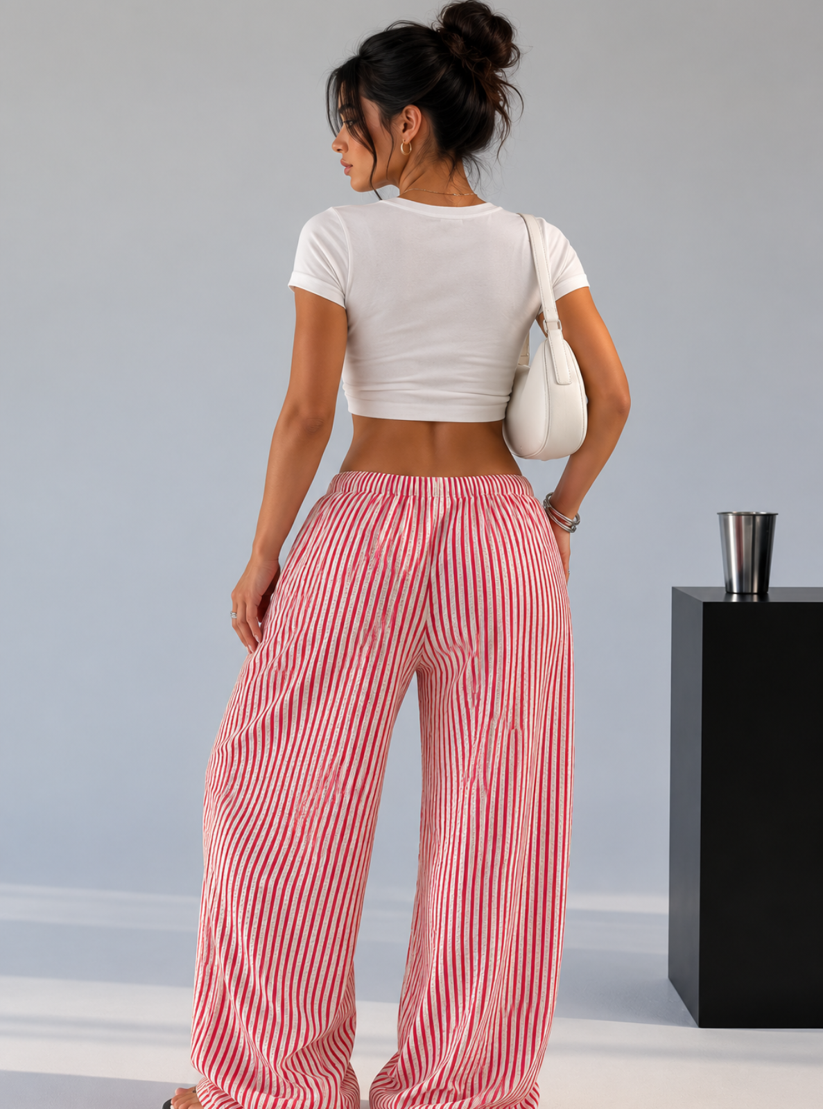 A woman shown from the back wearing pink and white vertically striped wide-leg pants, a white crop top, and carrying a white shoulder bag, standing in a studio setting.