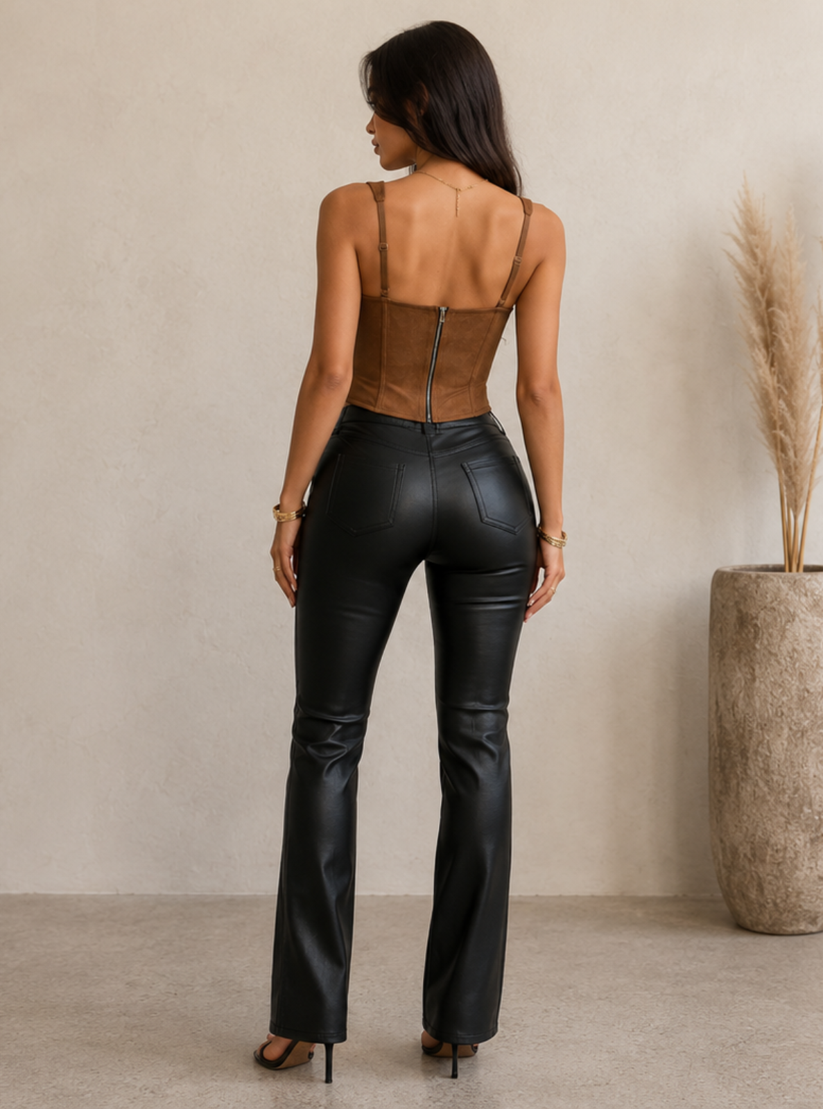 A woman stands with her back to the camera wearing a brown suede-like corset top and black faux-leather flare pants, posed in a minimalist room with a textured beige wall and a large vase of pampas grass.