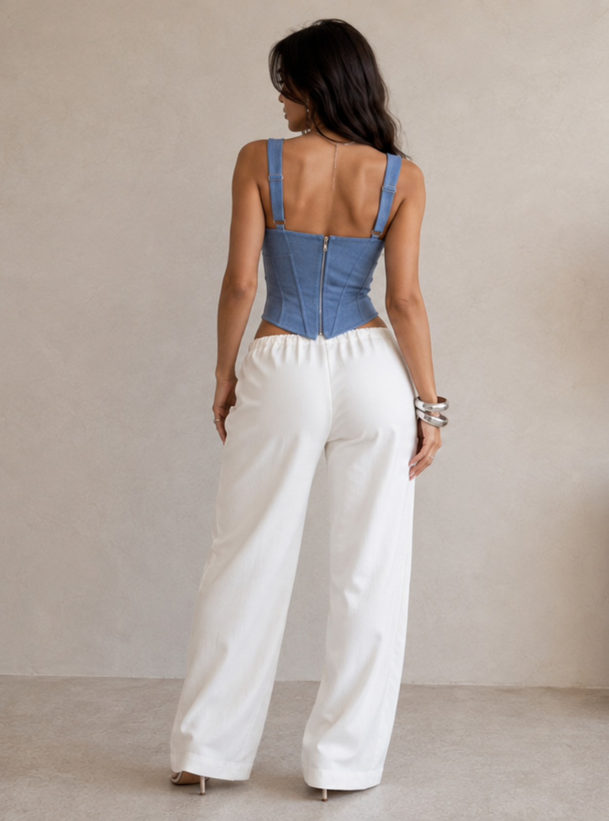 A woman stands with her back to the camera wearing a blue denim corset-style top and white wide-leg pants in a minimalist beige room with a large vase of dried grass.