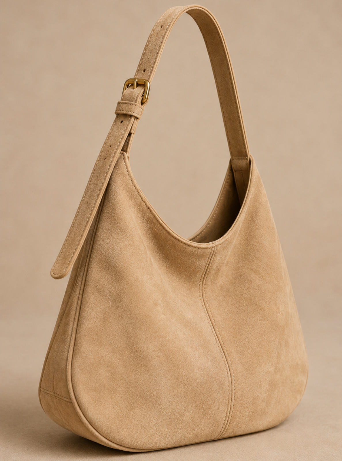 A beige suede hobo shoulder bag with a single adjustable strap and a gold-tone buckle, shown on a neutral beige background.