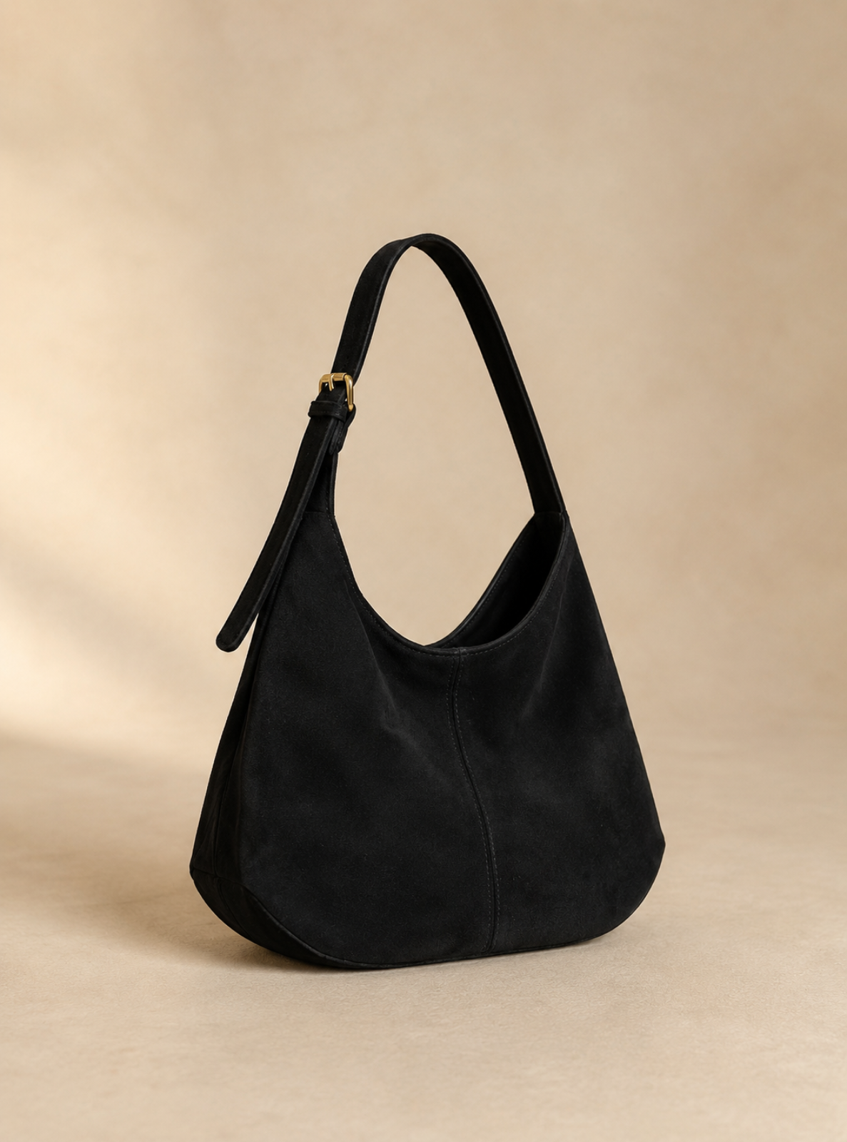 A black suede hobo shoulder bag with a single shoulder strap and a small gold-tone buckle, displayed on a warm beige studio background.