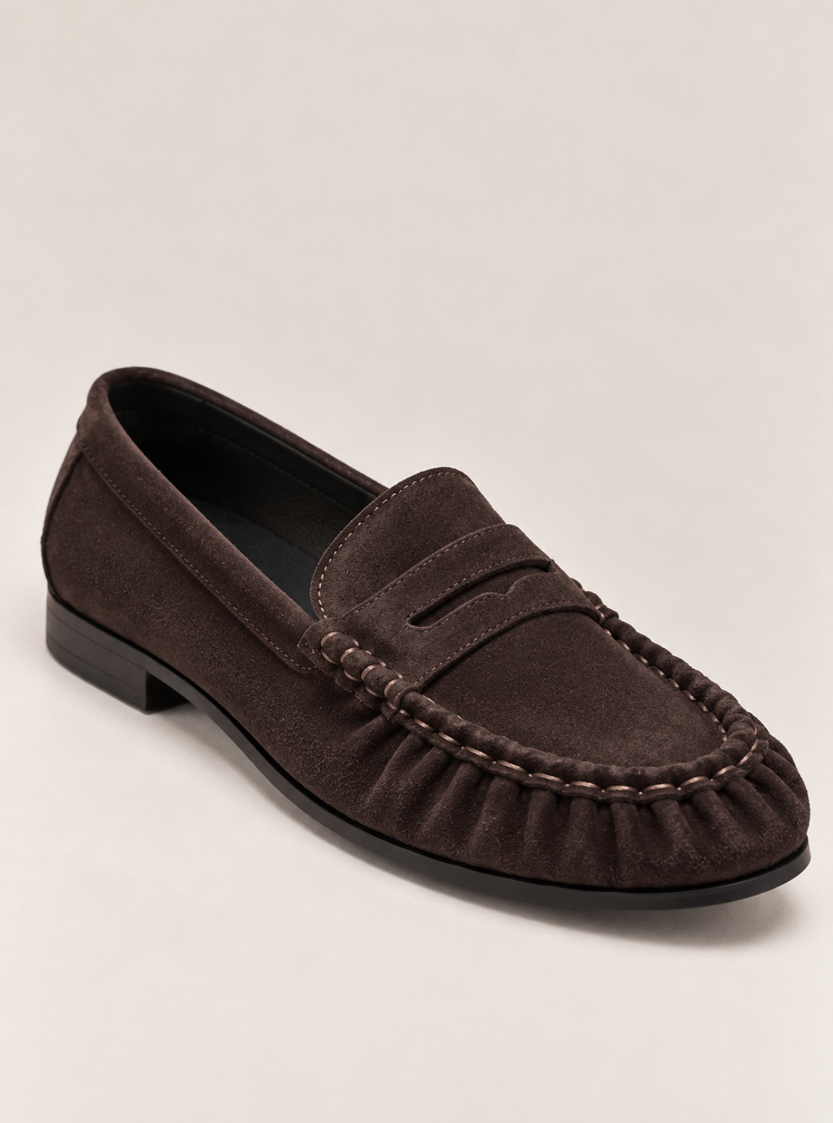 A dark brown suede penny loafer with decorative stitching and a low block heel, photographed on a light beige background.