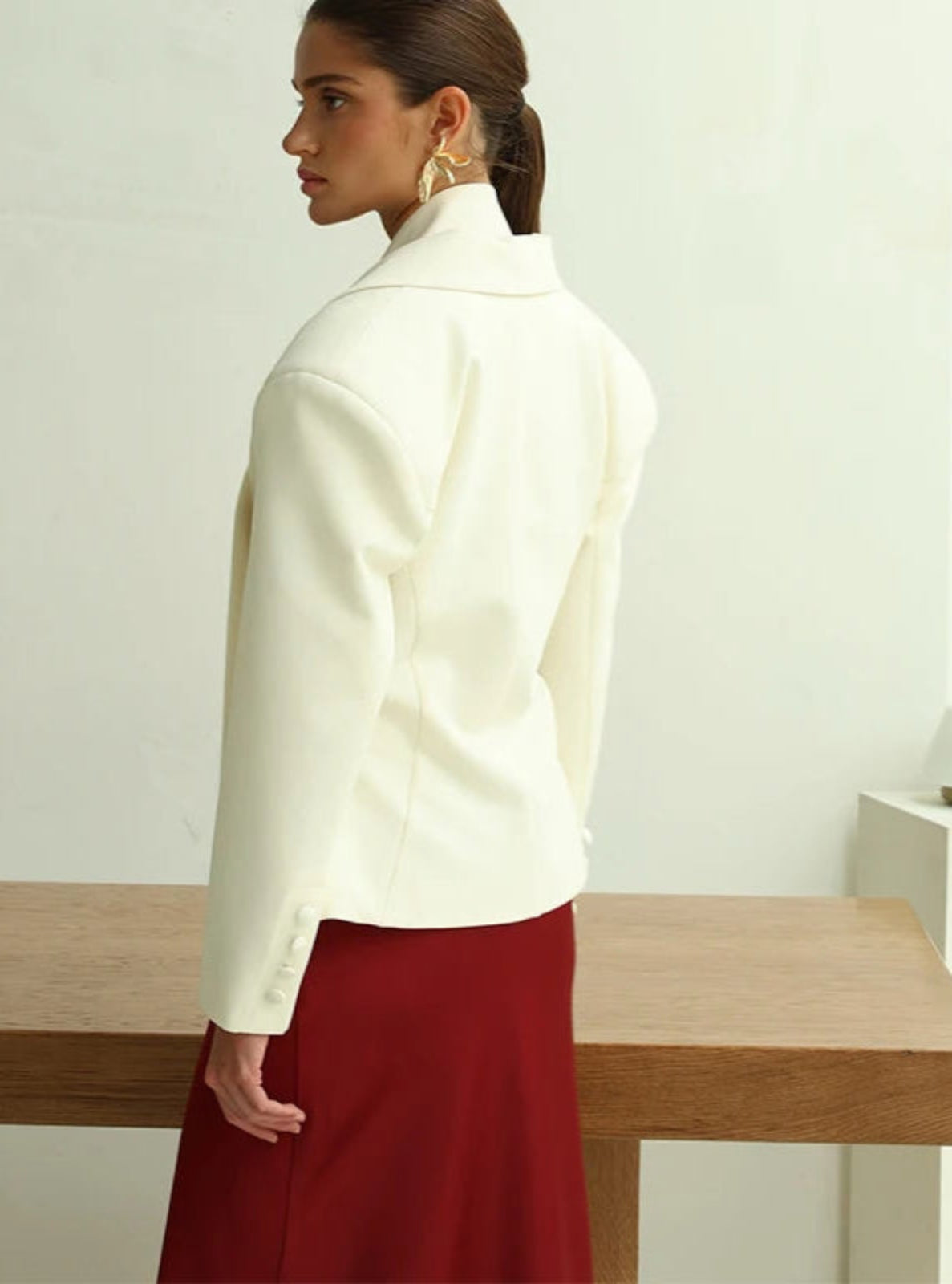Woman wearing a white jacket and red skirt standing in a minimalistic room.
