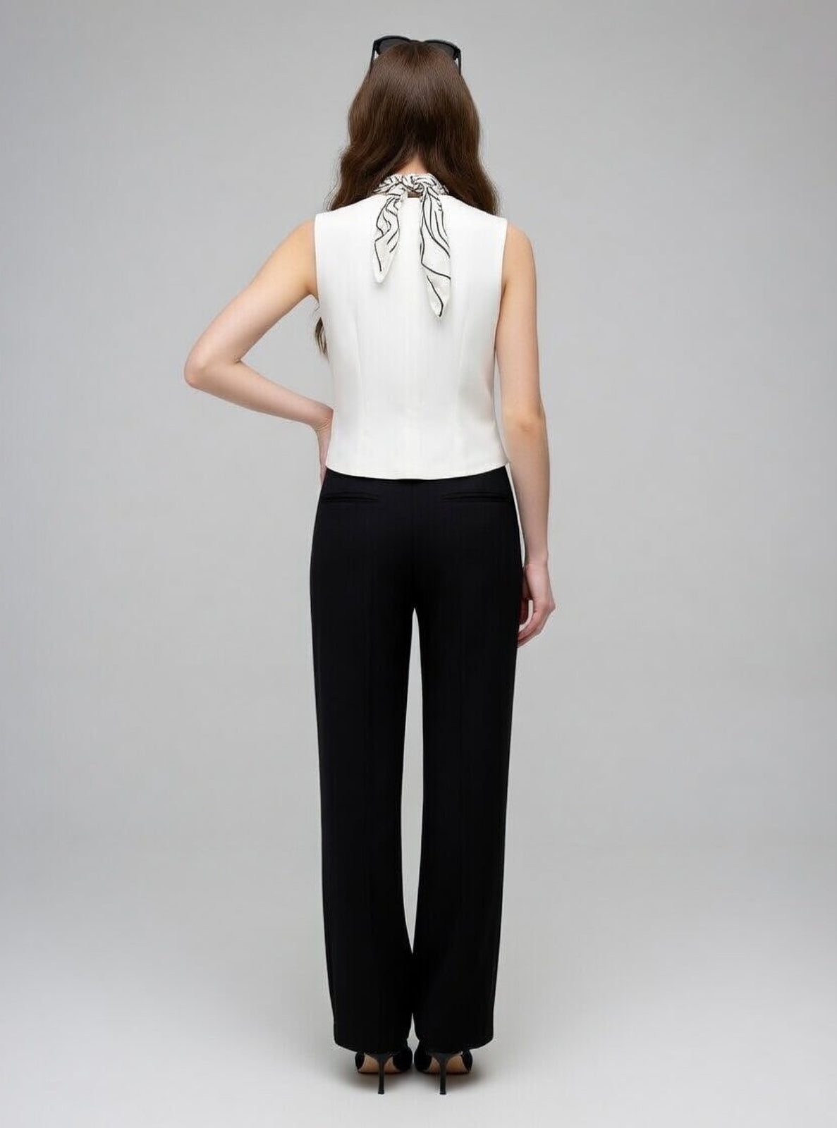 Woman wearing a white sleeveless top with a decorative back and black pants on a gray background