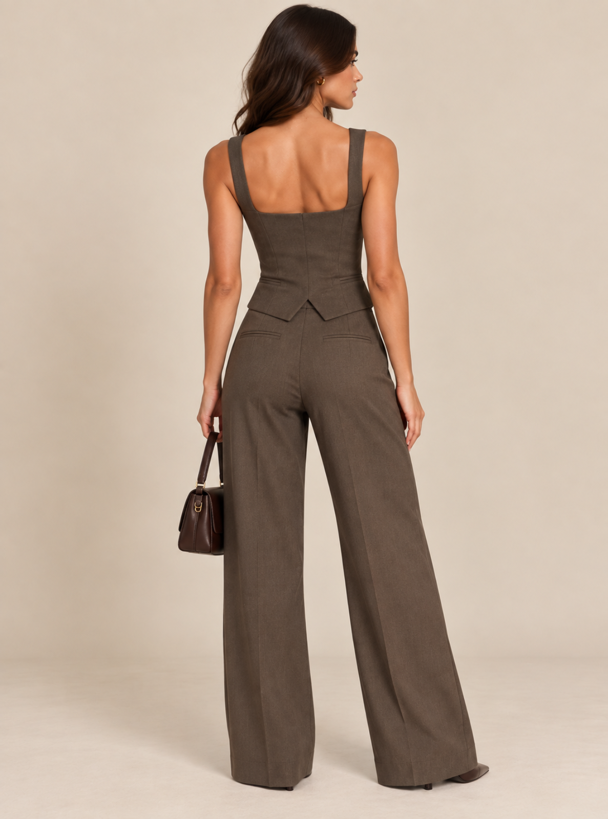 A woman shown from the back wearing a dark brown wide-leg jumpsuit with a structured bodice and shoulder straps, holding a small dark brown handbag against a plain beige studio background.