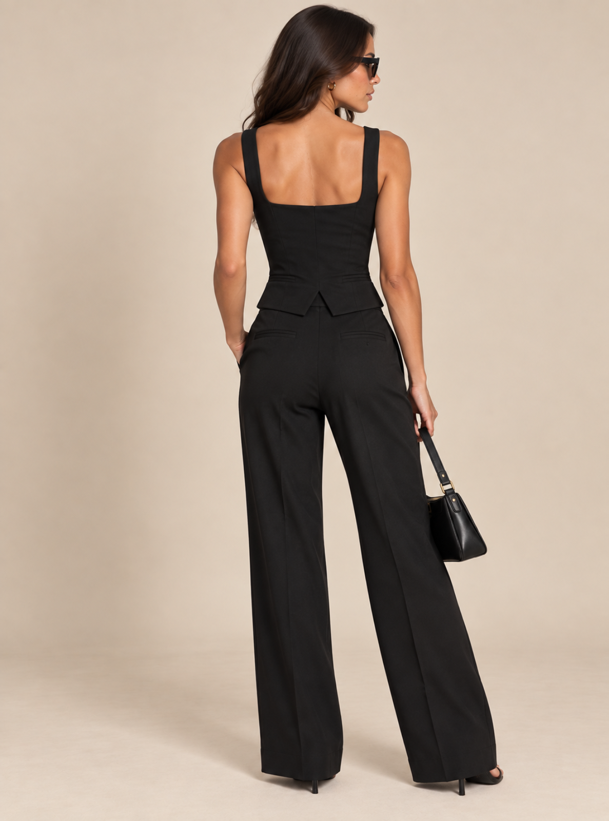 A woman photographed from the back wearing a black sleeveless wide-leg jumpsuit with a structured peplum waist, paired with black high heels and a small black handbag, against a plain beige studio background.
