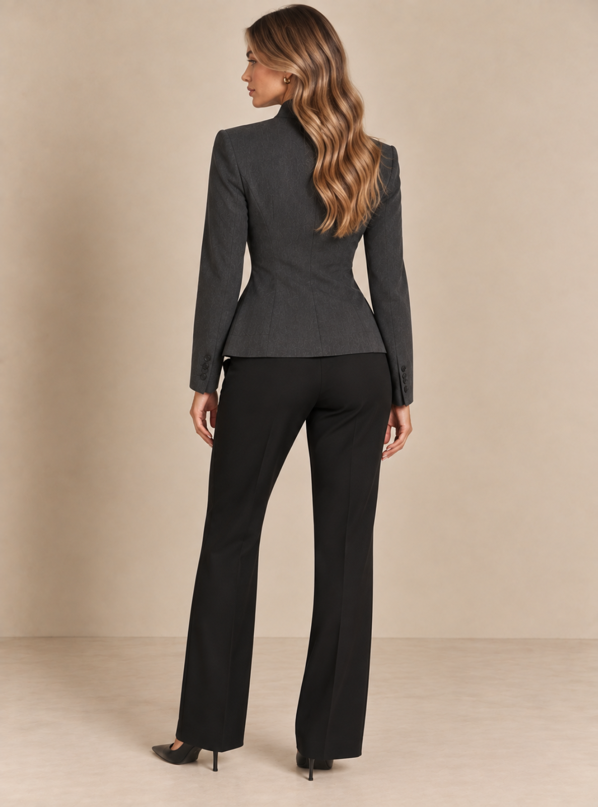 A woman shown from the back wearing a dark gray structured blazer and black wide-leg trousers, standing against a plain beige studio backdrop.