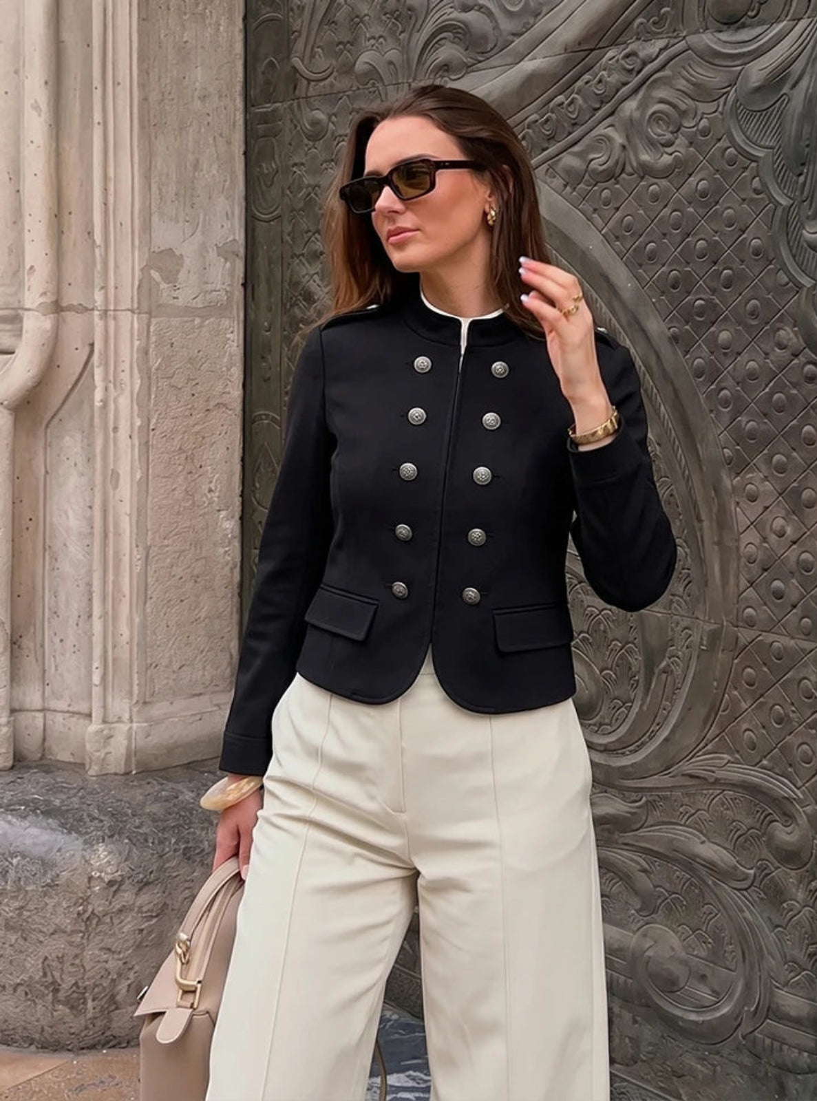 Woman wearing a black jacket with silver buttons and white pants, standing against a textured stone wall.