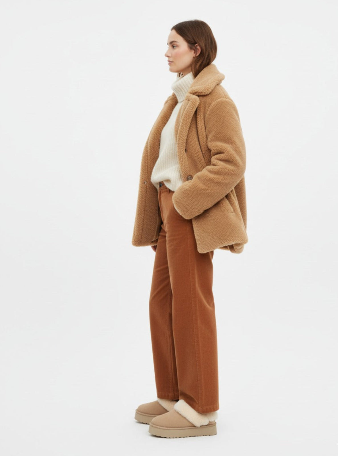 Woman wearing a brown coat, white sweater, and brown pants on a white background