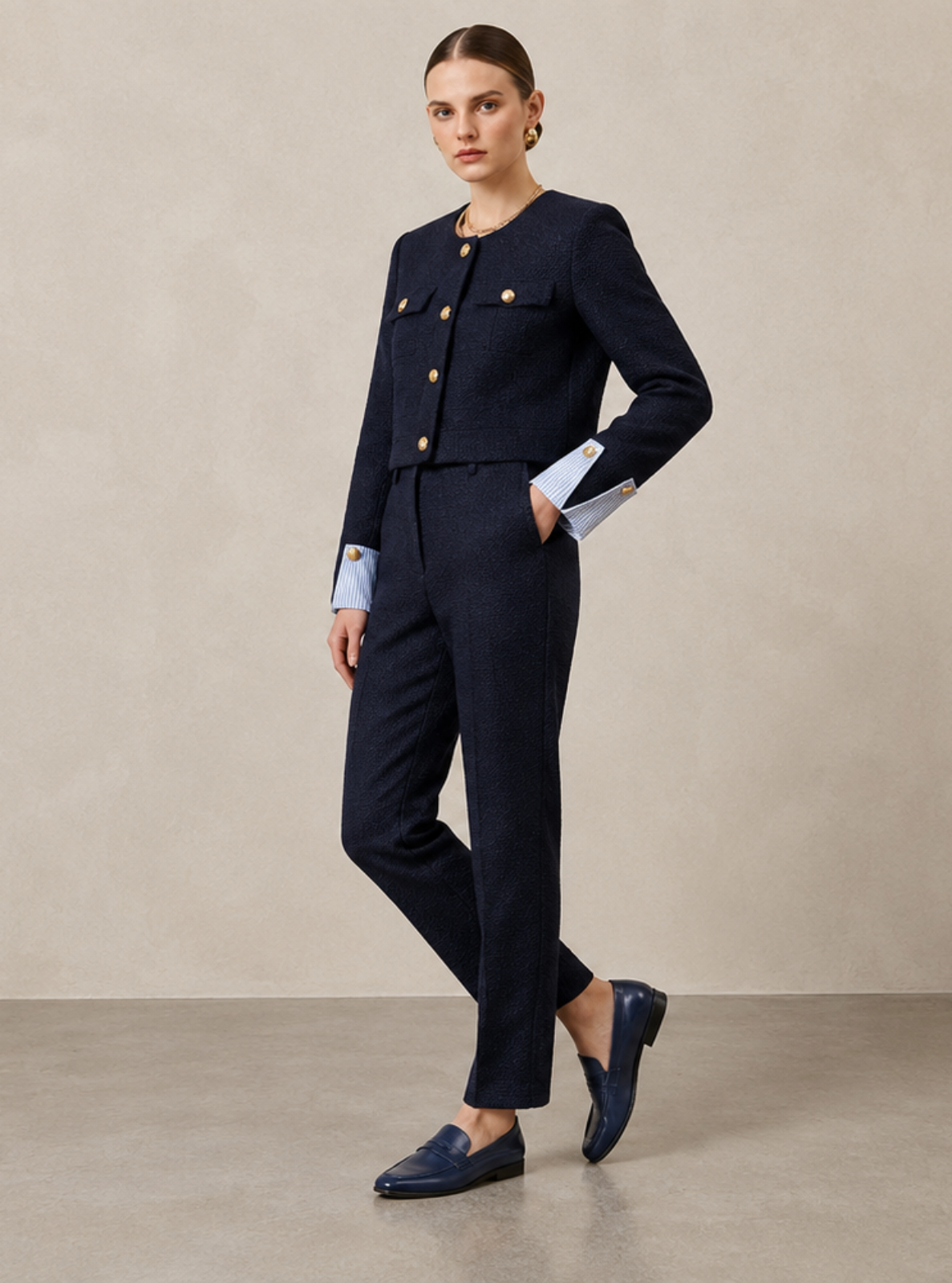 A woman models a dark navy blue tailored set consisting of a cropped jacket with gold buttons and matching trousers, styled with a light blue shirt at the cuffs and navy loafers, in a minimalist studio setting.