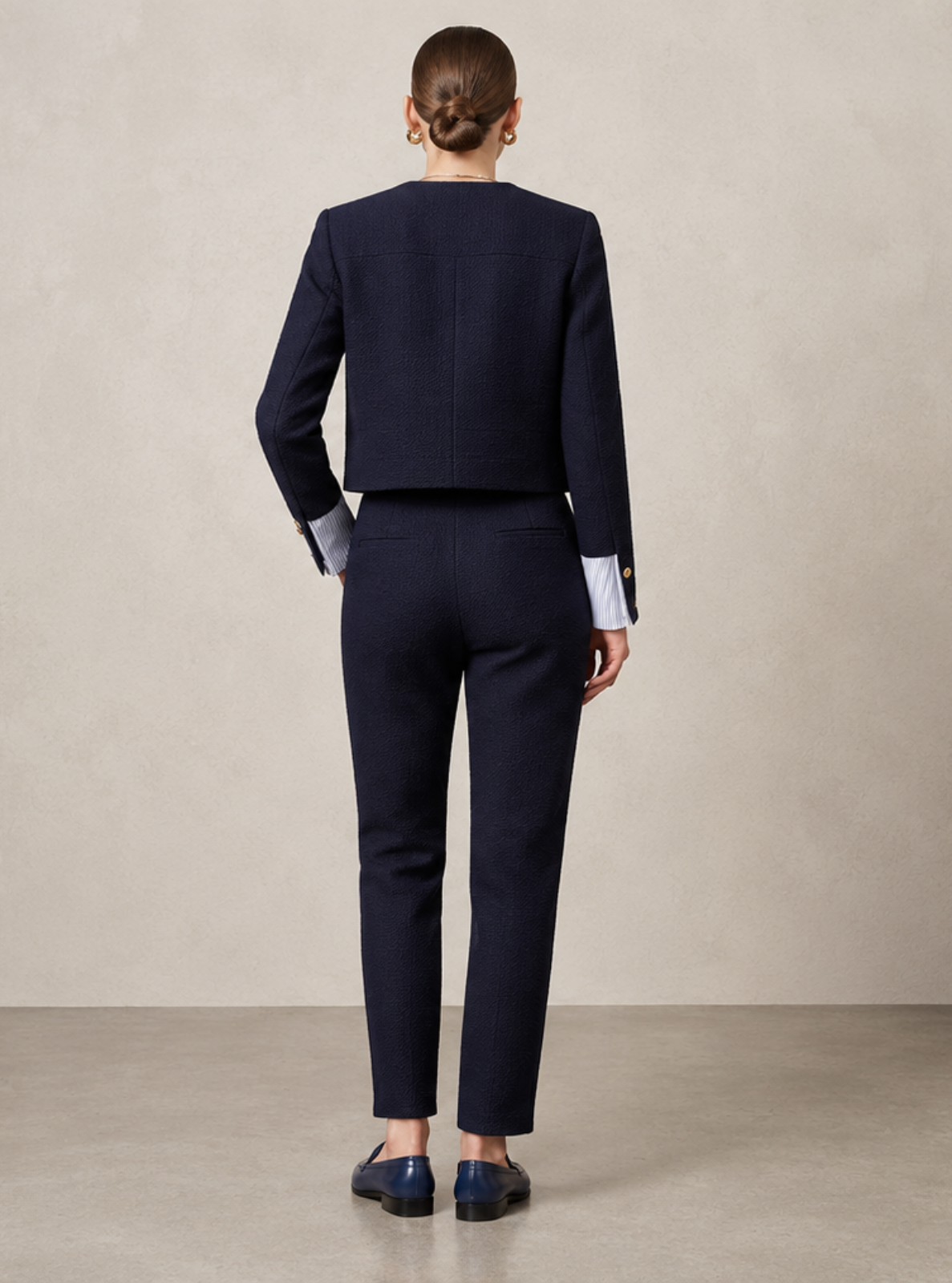 A model viewed from behind wearing a dark navy blue tailored suit with a cropped jacket and straight-leg trousers, standing in a minimalist studio with a beige textured wall and a small wooden stool.