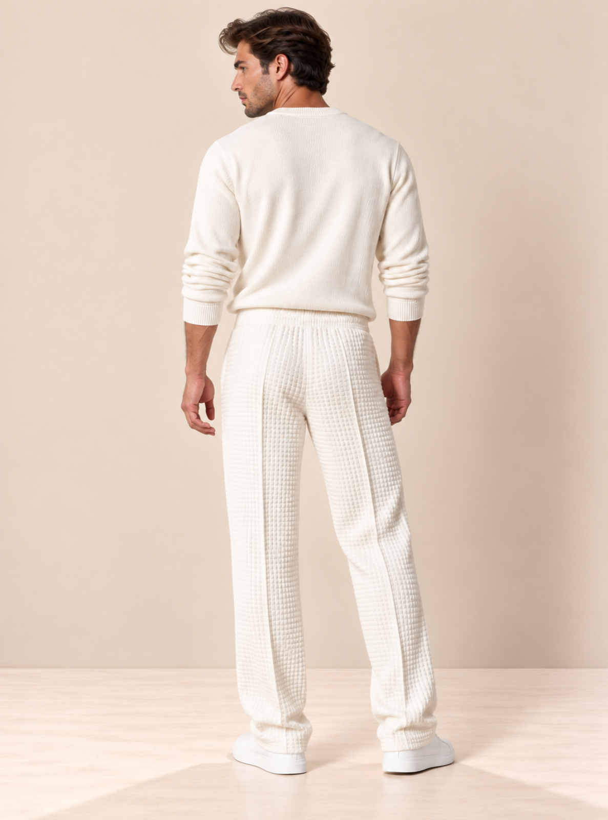 A man shown from the back wearing a cream-colored knit sweater and matching textured knit pants against a warm beige studio background.