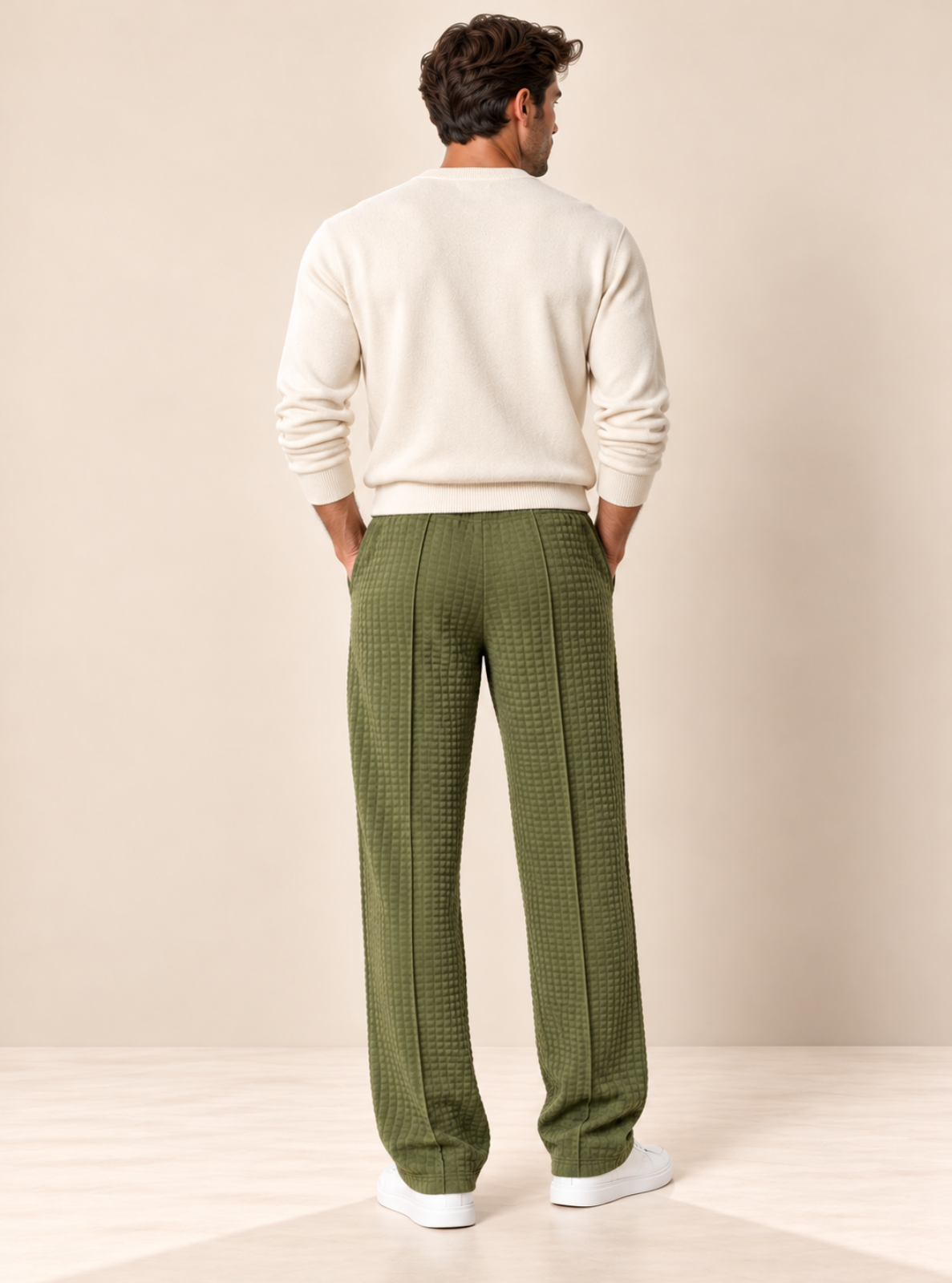 A man is shown from behind wearing olive green textured trousers, a cream knit sweater, and white sneakers, standing against a warm beige studio backdrop.