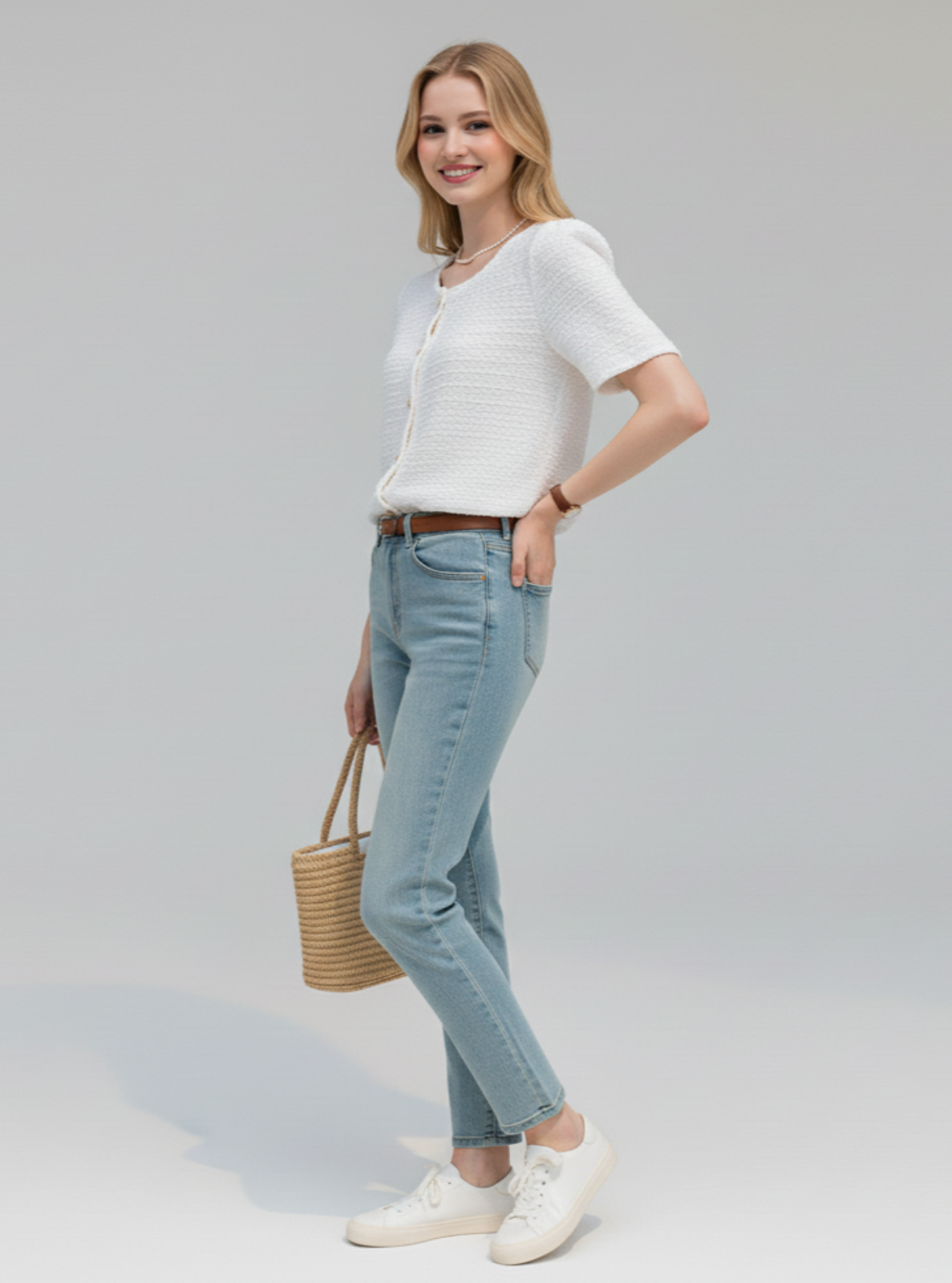 Woman wearing a white top and light blue jeans, holding a woven bag on a plain background