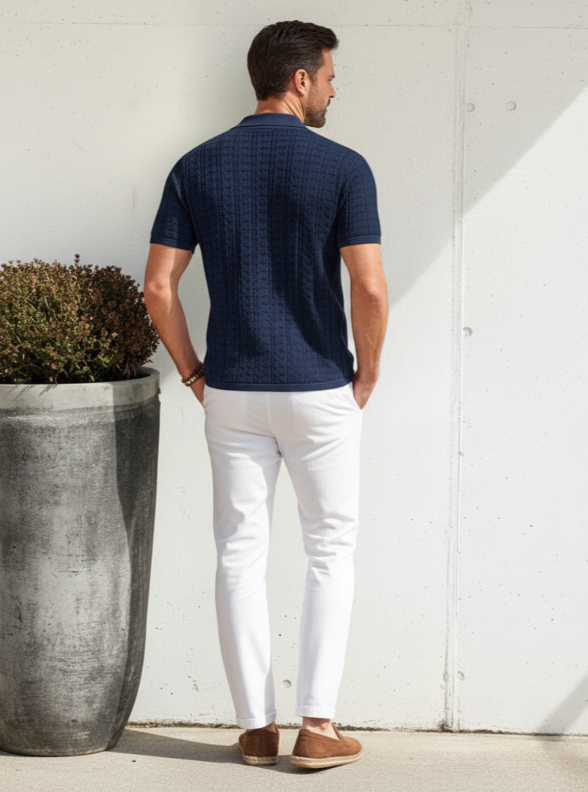 Man in navy shirt and white pants standing next to a large planter against a light-colored wall.