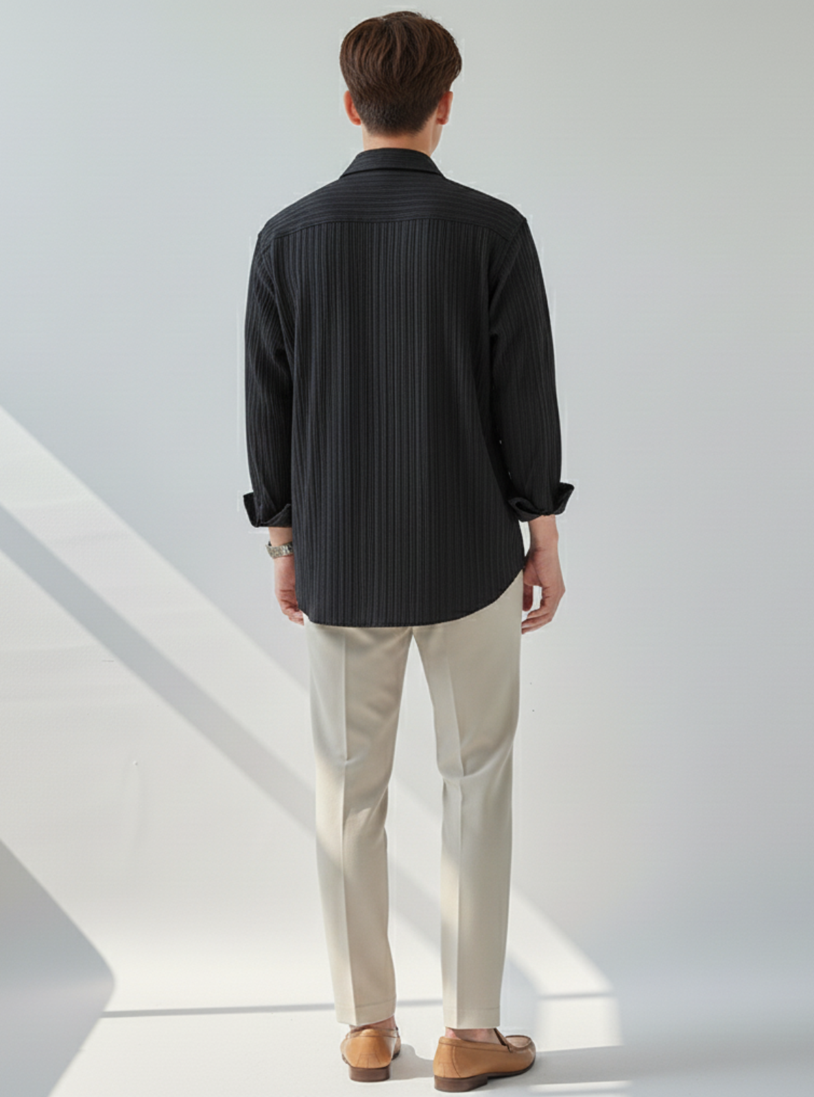 Man wearing a black pinstripe shirt and beige pants standing in a minimalistic setting.