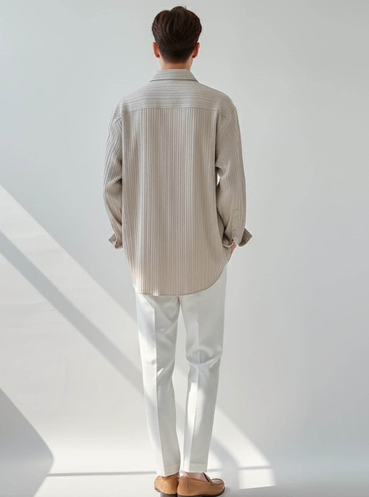 Person wearing a striped shirt and white pants standing in a minimalistic room with sunlight casting shadows.