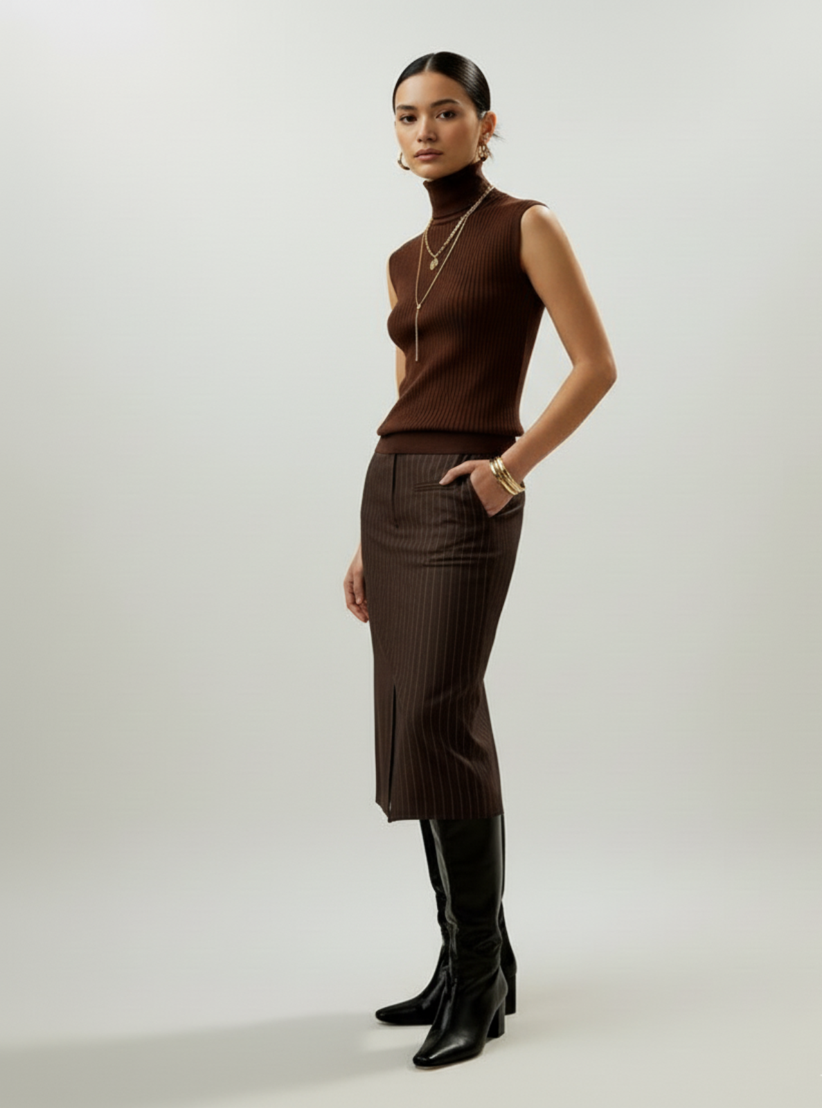 Woman wearing a brown sleeveless top and pinstripe skirt with black knee-high boots on a white background