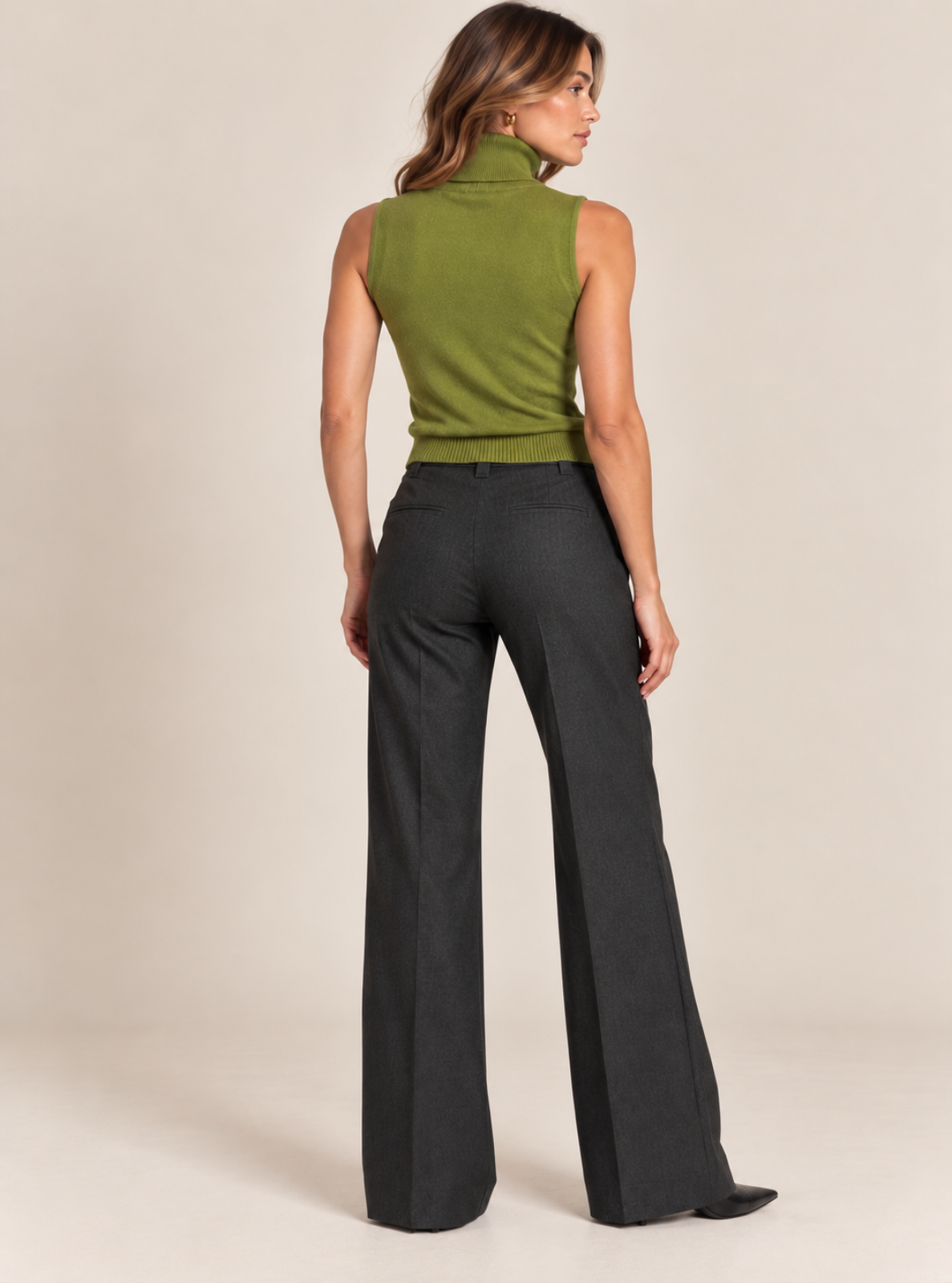 A woman shown from the back wearing a sleeveless olive green mock-neck top and dark charcoal gray wide-leg trousers, standing against a plain beige studio background.