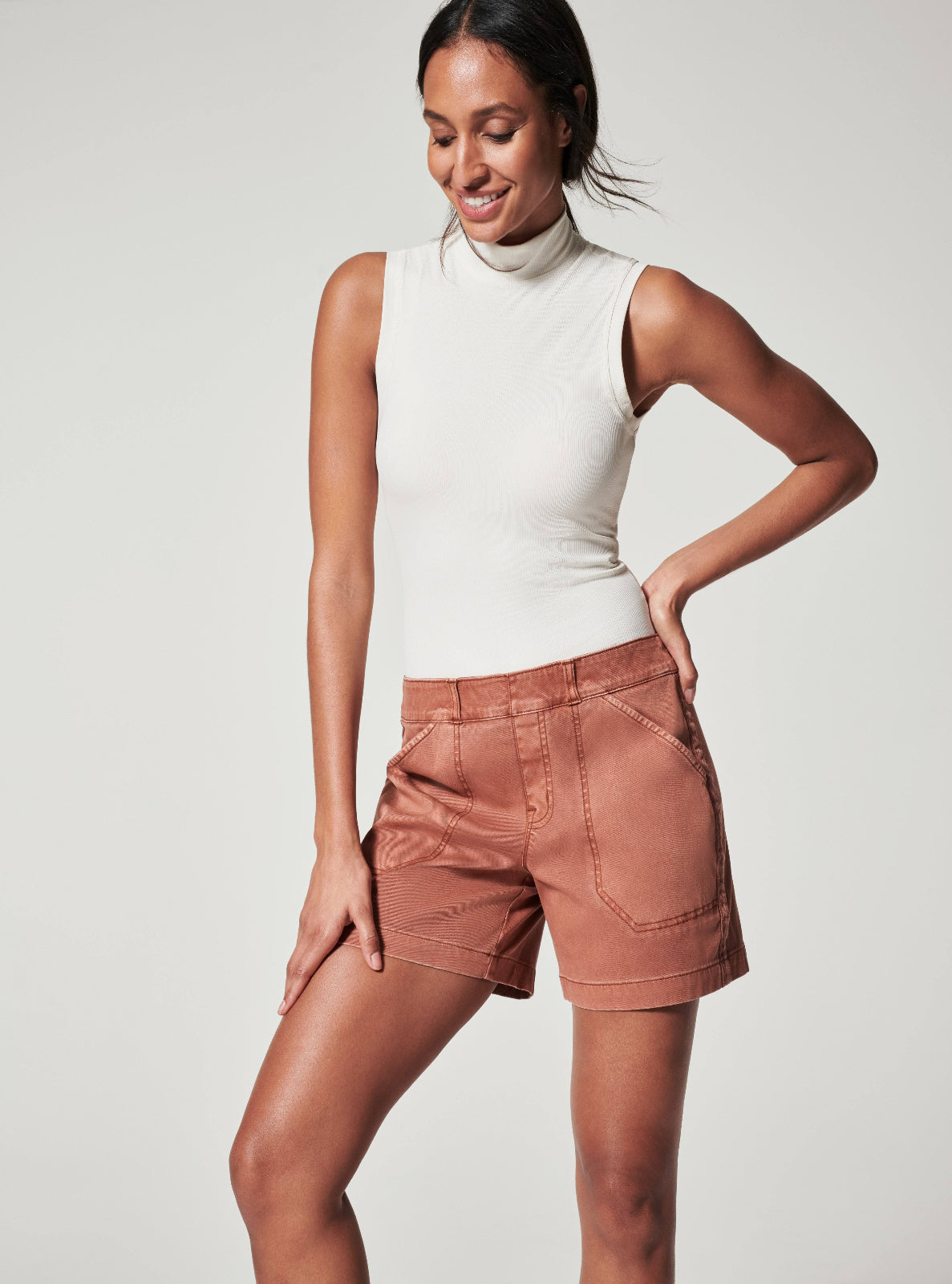 Woman wearing a white sleeveless top and brown shorts on a light gray background