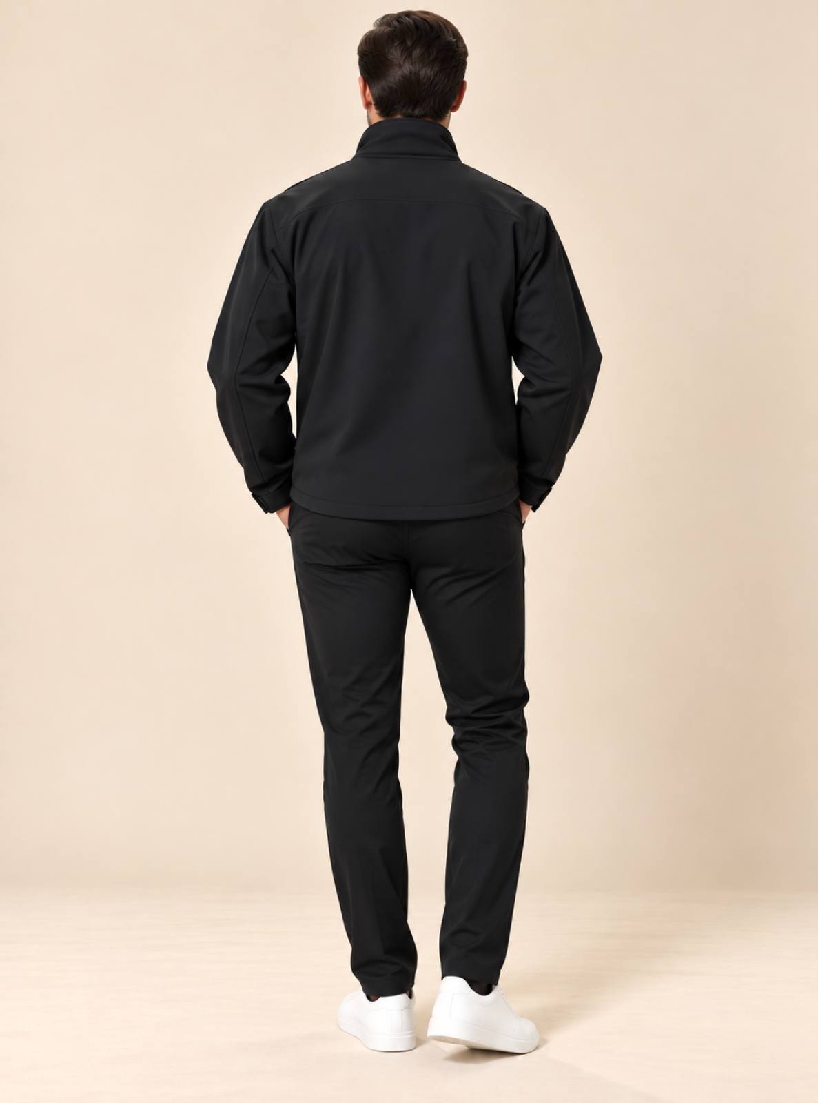 A man viewed from behind wearing a black zip-up jacket and black pants, standing against a plain beige studio background.