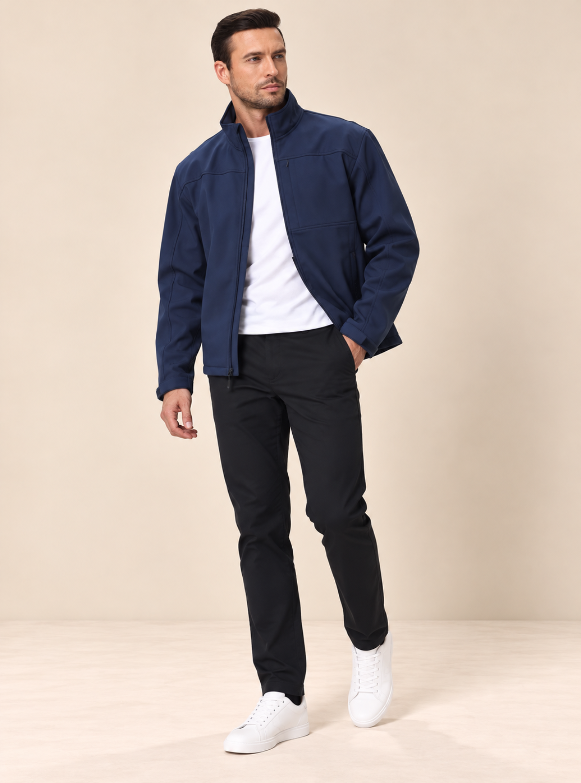 A man modeling a navy blue lightweight jacket over a white t-shirt, black trousers, and white sneakers, standing against a solid beige studio background.