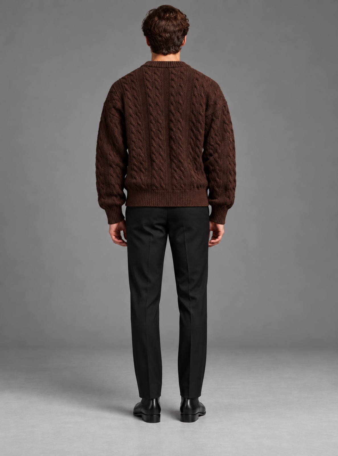 A man viewed from the back wearing a dark brown cable-knit sweater, black trousers, and black shoes, standing against a neutral gray studio background.