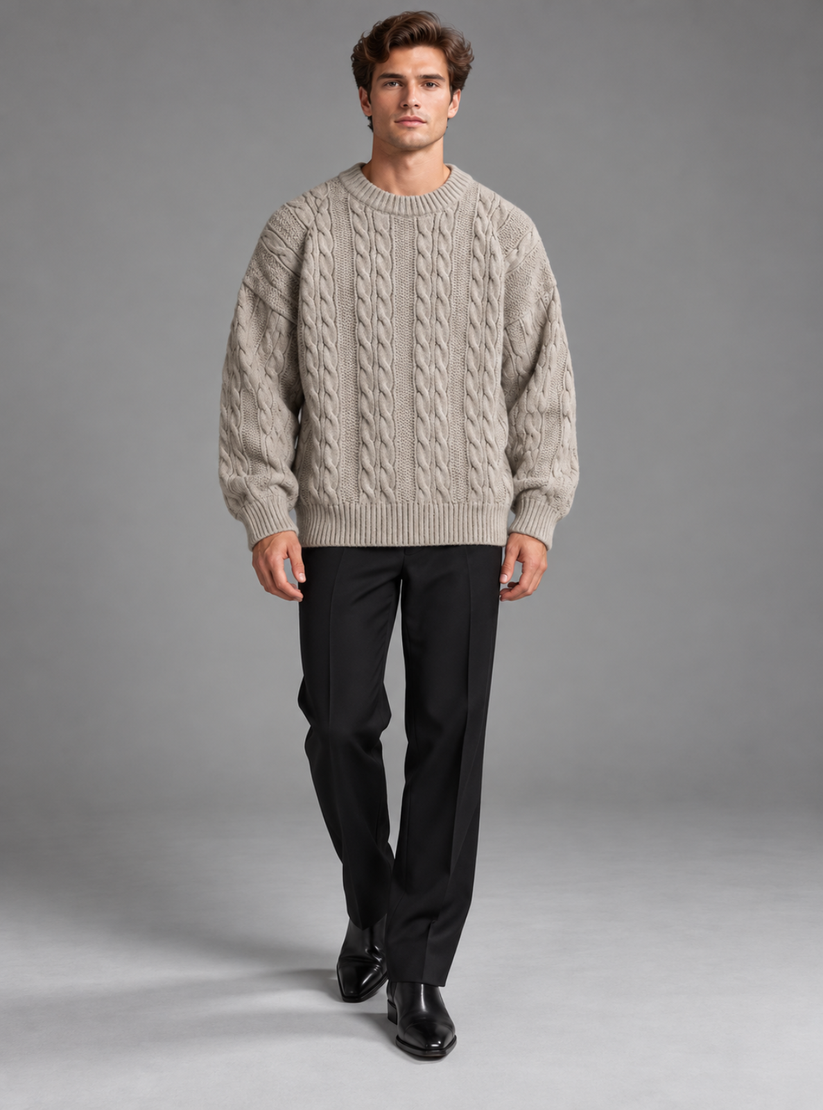 A man modeling a light beige cable-knit crewneck sweater, paired with black trousers and black leather shoes, against a solid gray studio background.