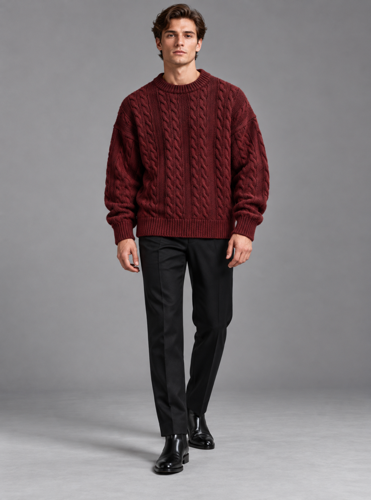 A man modeling a burgundy cable-knit crewneck sweater, paired with black trousers and black leather shoes, against a solid gray studio background.