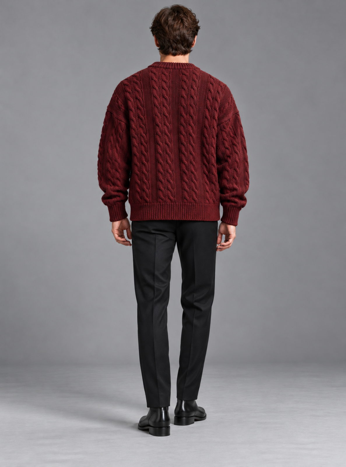 A man viewed from behind wearing a burgundy cable-knit sweater, black trousers, and black shoes, standing against a neutral gray studio background.