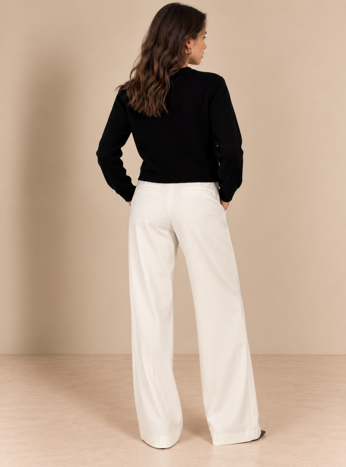 A woman shown from the back wearing a black long-sleeve top and off-white wide-leg trousers, standing against a solid beige studio background.