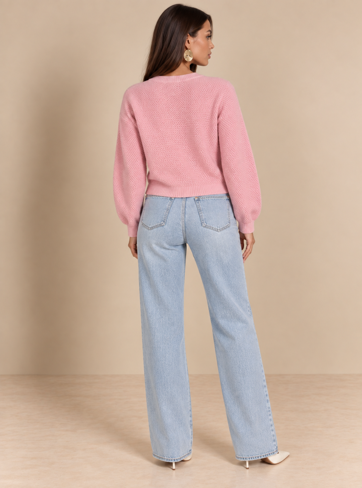 A woman is shown from the back wearing a light pink knit sweater and light blue wide-leg jeans, standing against a solid beige studio background.