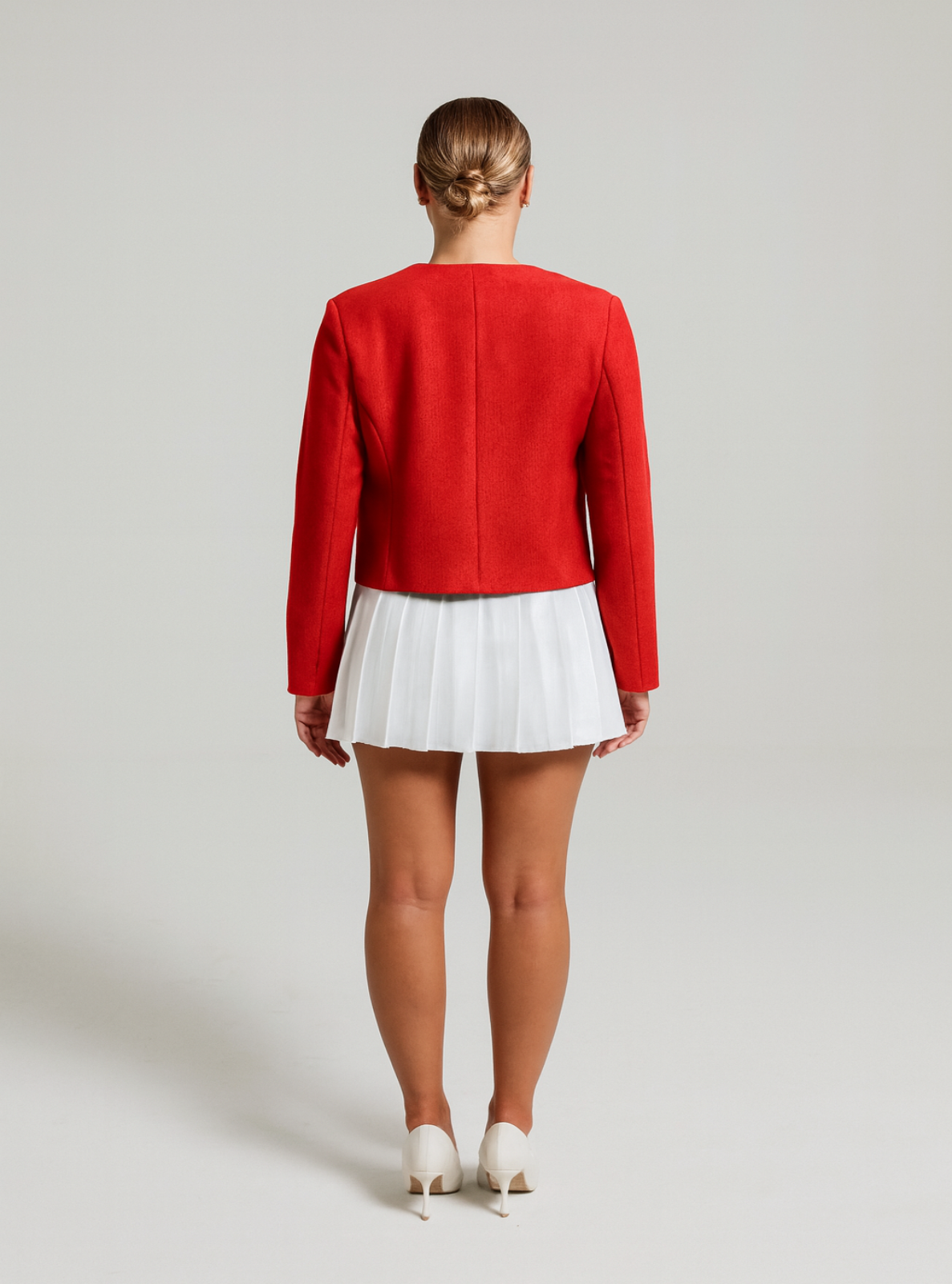 Model in a sleek ponytail wearing a red textured Jacket with gold buttons, a white pleated skirt facing the white background.
