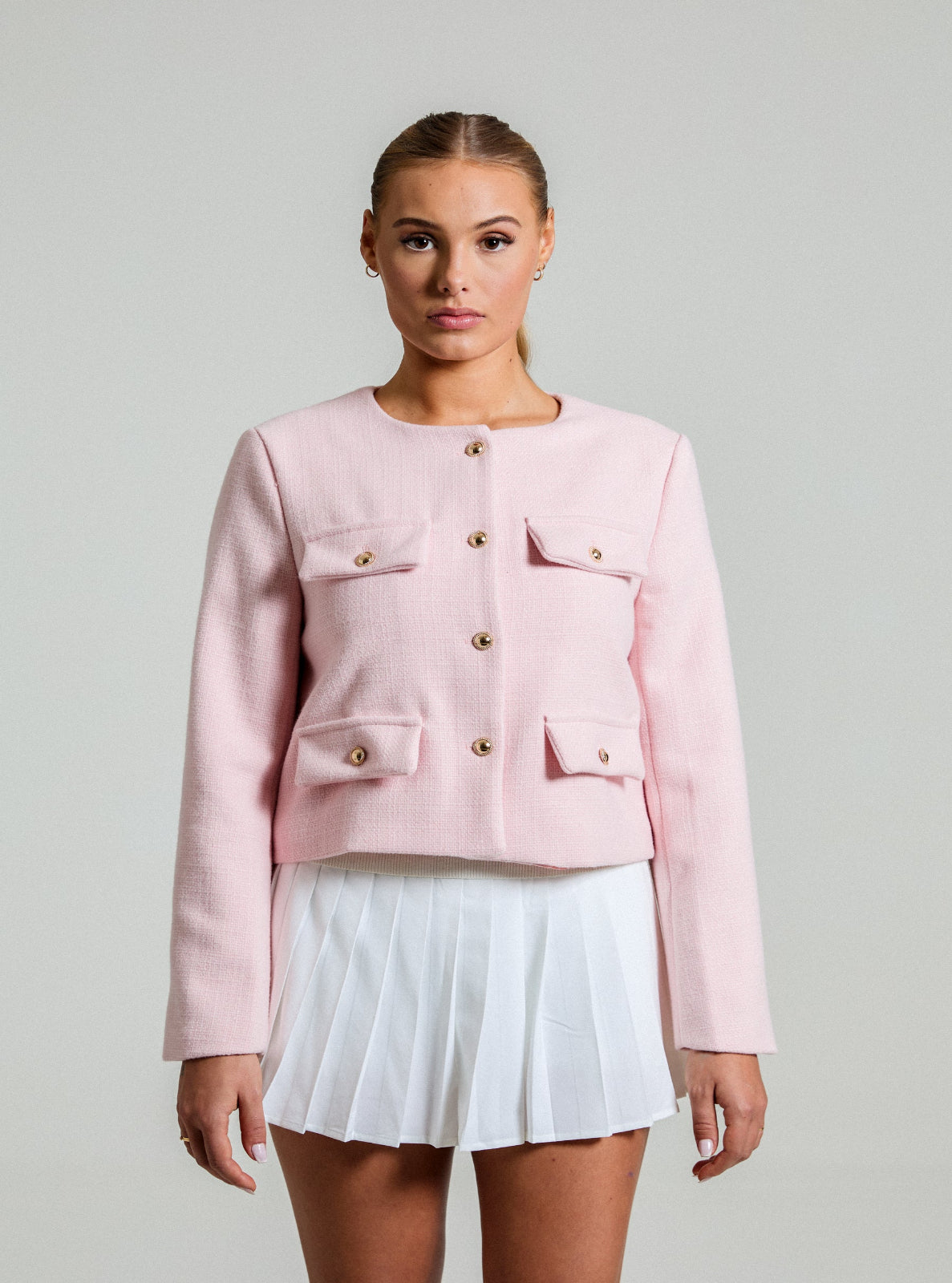 model wearing a a pink textured Jacket with gold buttons and a white tennis skirt in front of a white background