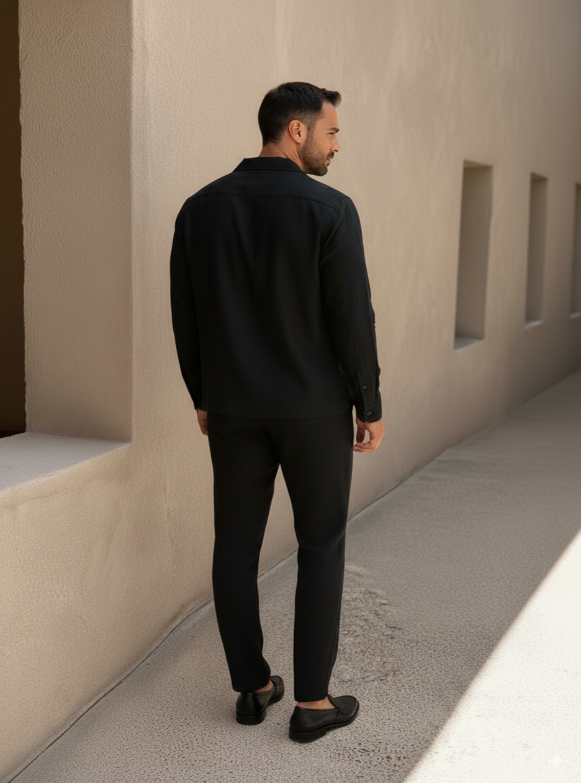 Man in a black outfit standing against a beige wall.