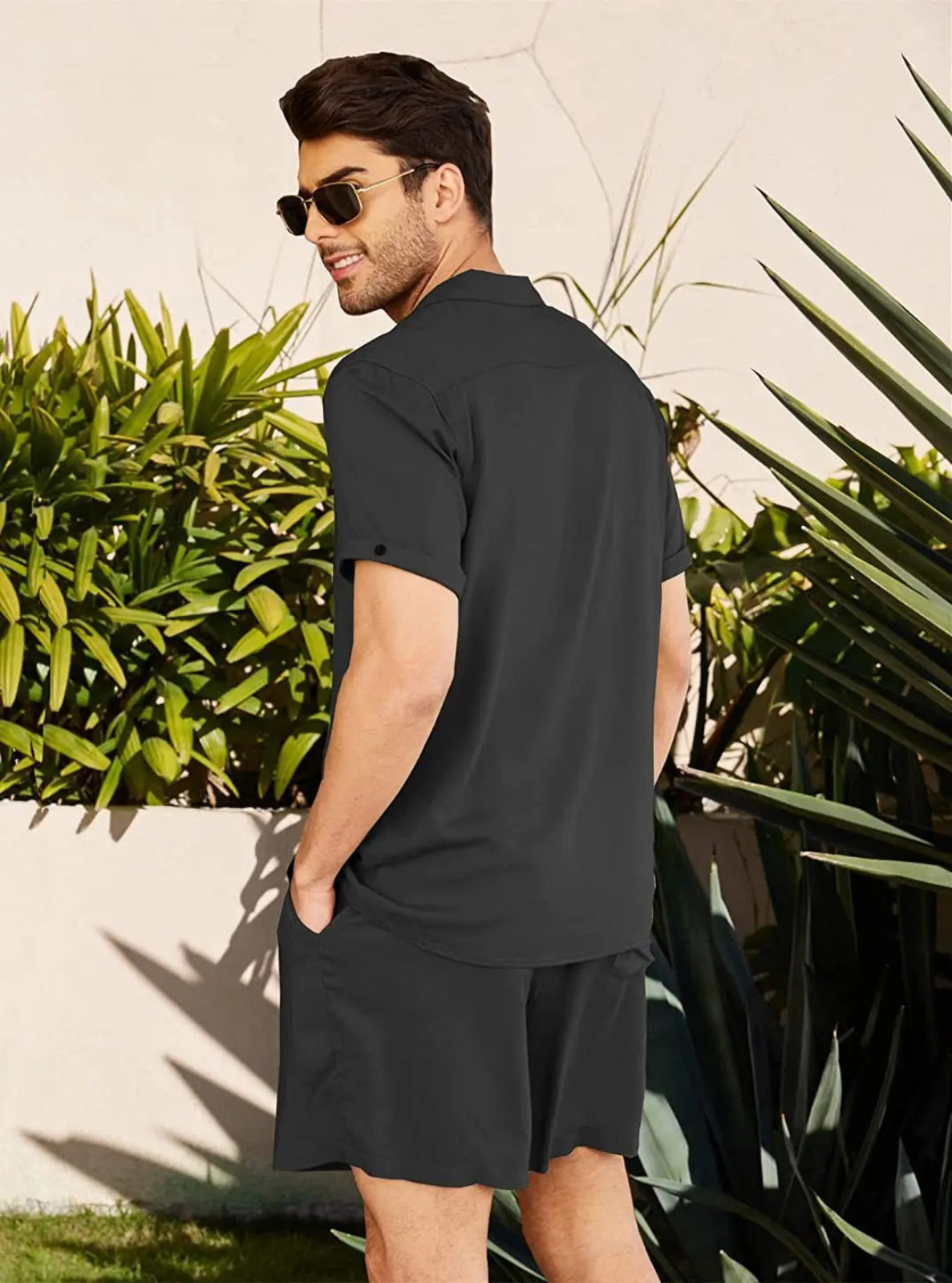 Man wearing a black polo shirt and shorts standing outdoors with plants in the background