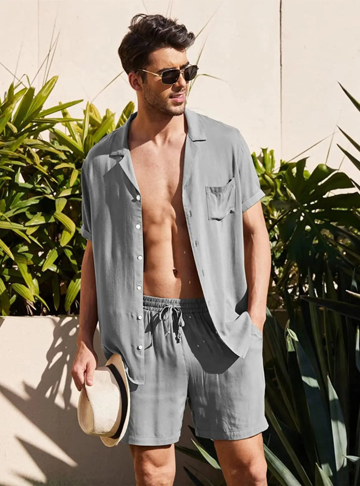 Man wearing a light gray shirt and shorts holding a hat, standing outdoors with plants in the background.
