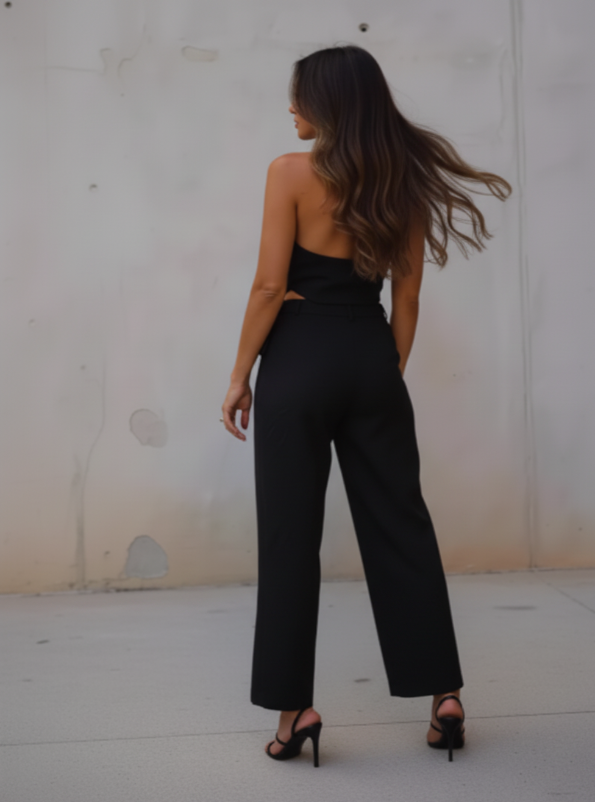 Woman wearing a black jumpsuit standing against a plain wall.