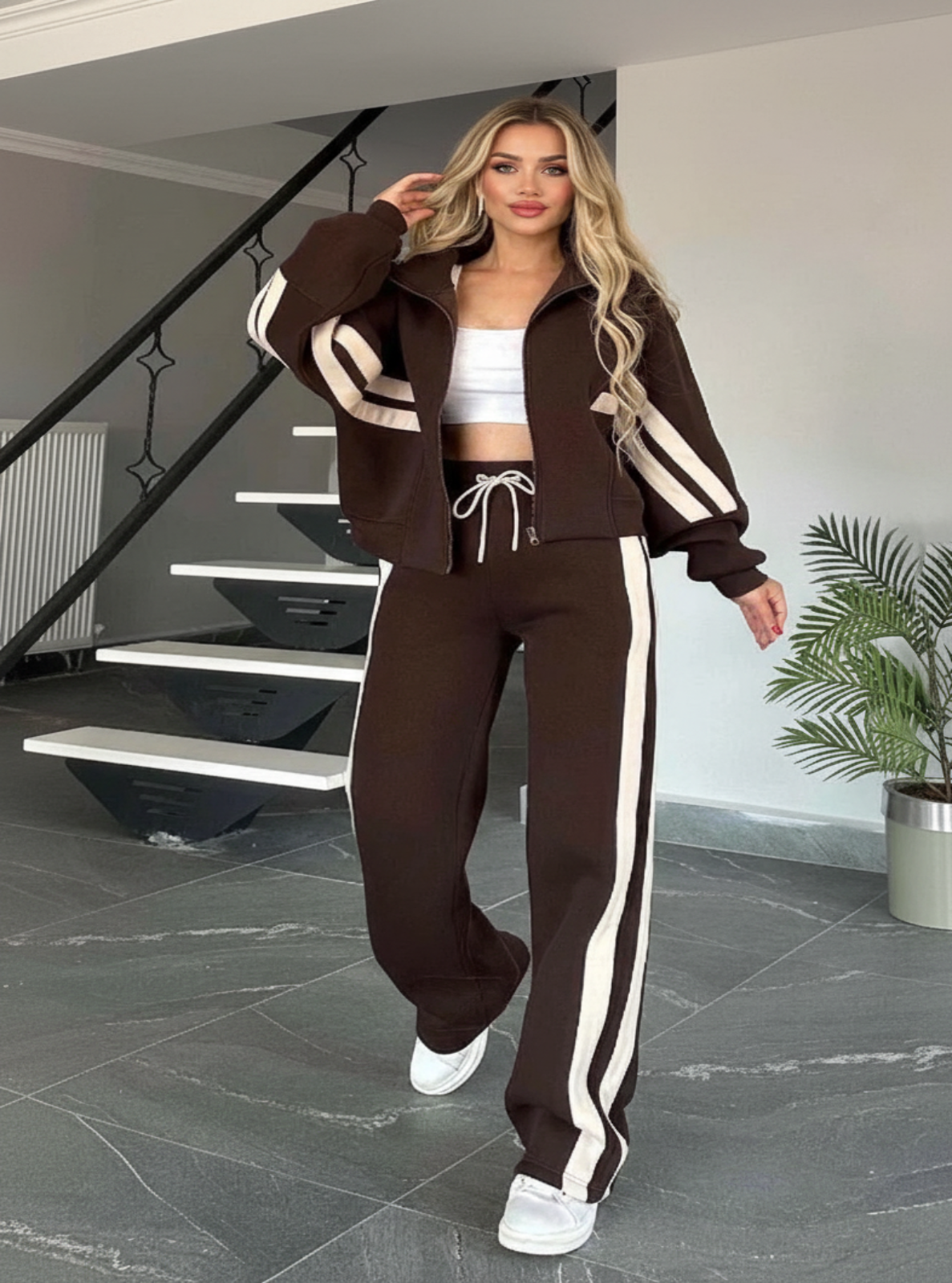 Woman in a brown tracksuit with white stripes standing in a modern interior setting.