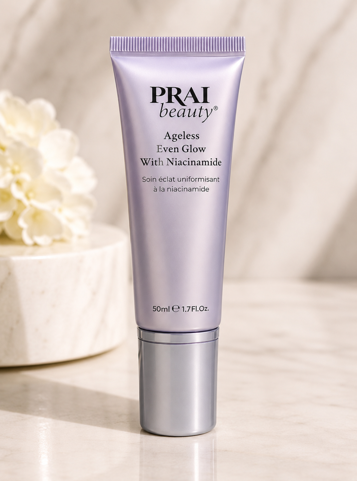 A tube of PRAI beauty Ageless Even Glow With Niacinamide skincare cream, standing upright on a light marble surface with a soft-focus floral background.