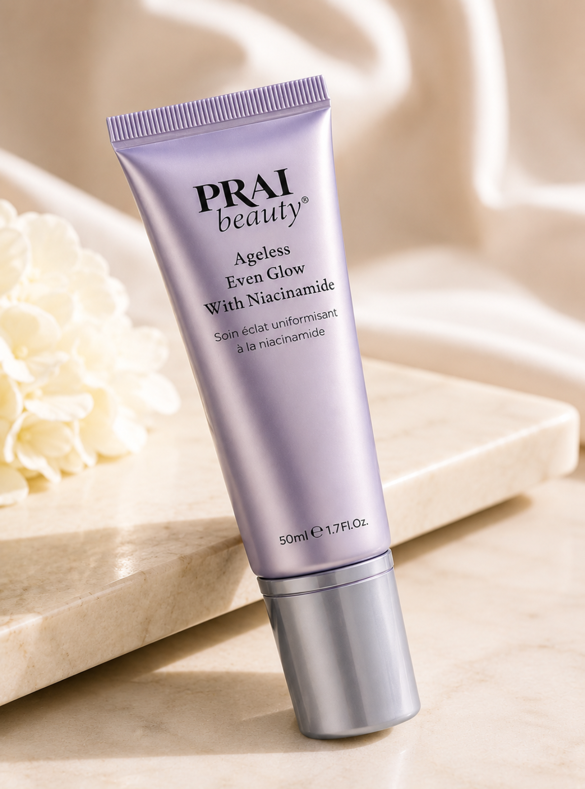 A lavender-purple squeeze tube of PRAI beauty Ageless Even Glow With Niacinamide, standing upright on a beige marble surface with soft cream-colored fabric and flowers in the background.