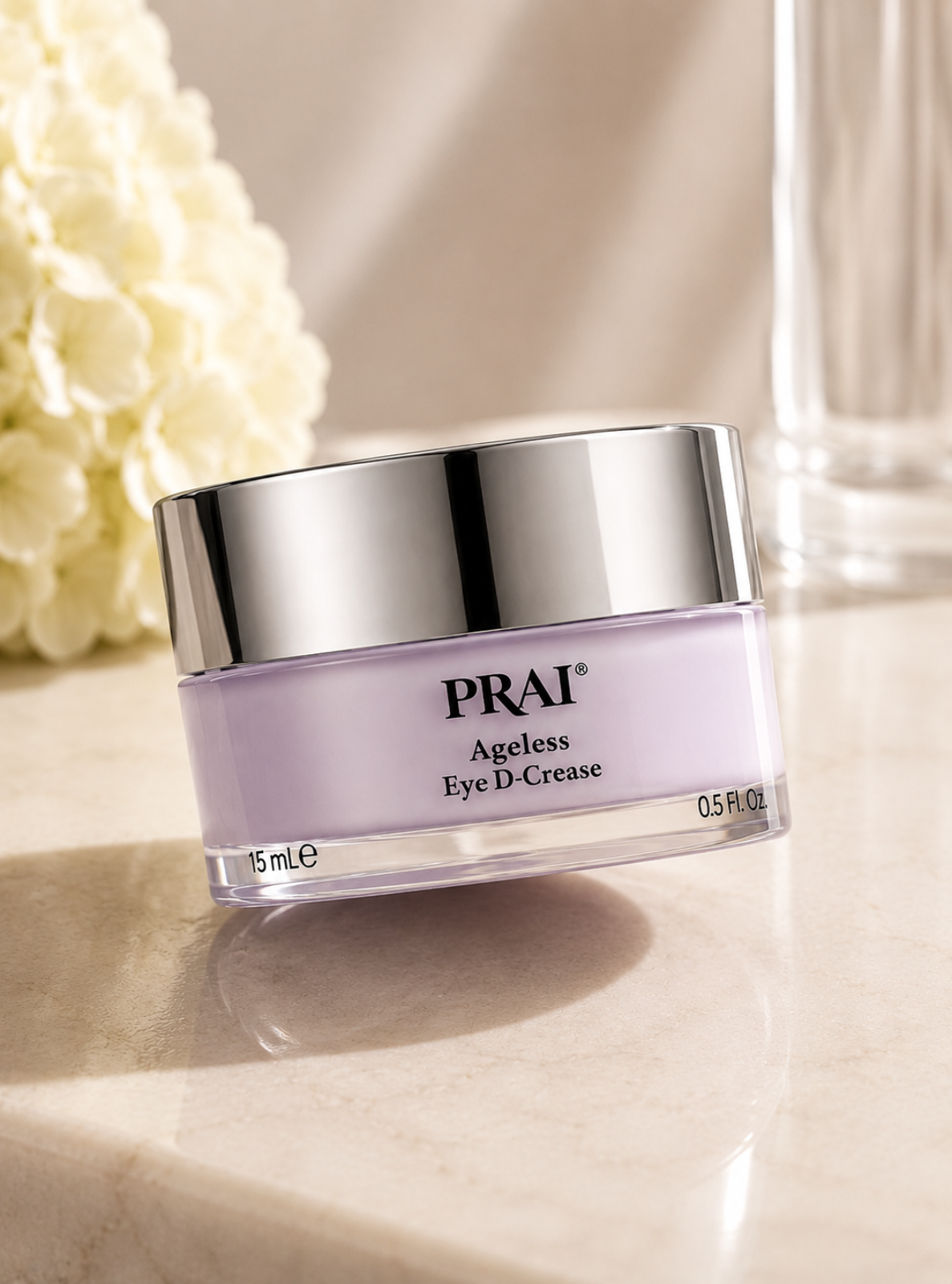 A jar of PRAI Ageless Eye D-Crease cream with a lavender-colored body and a shiny silver lid, set on a light marble surface with soft-focus flowers in the background.