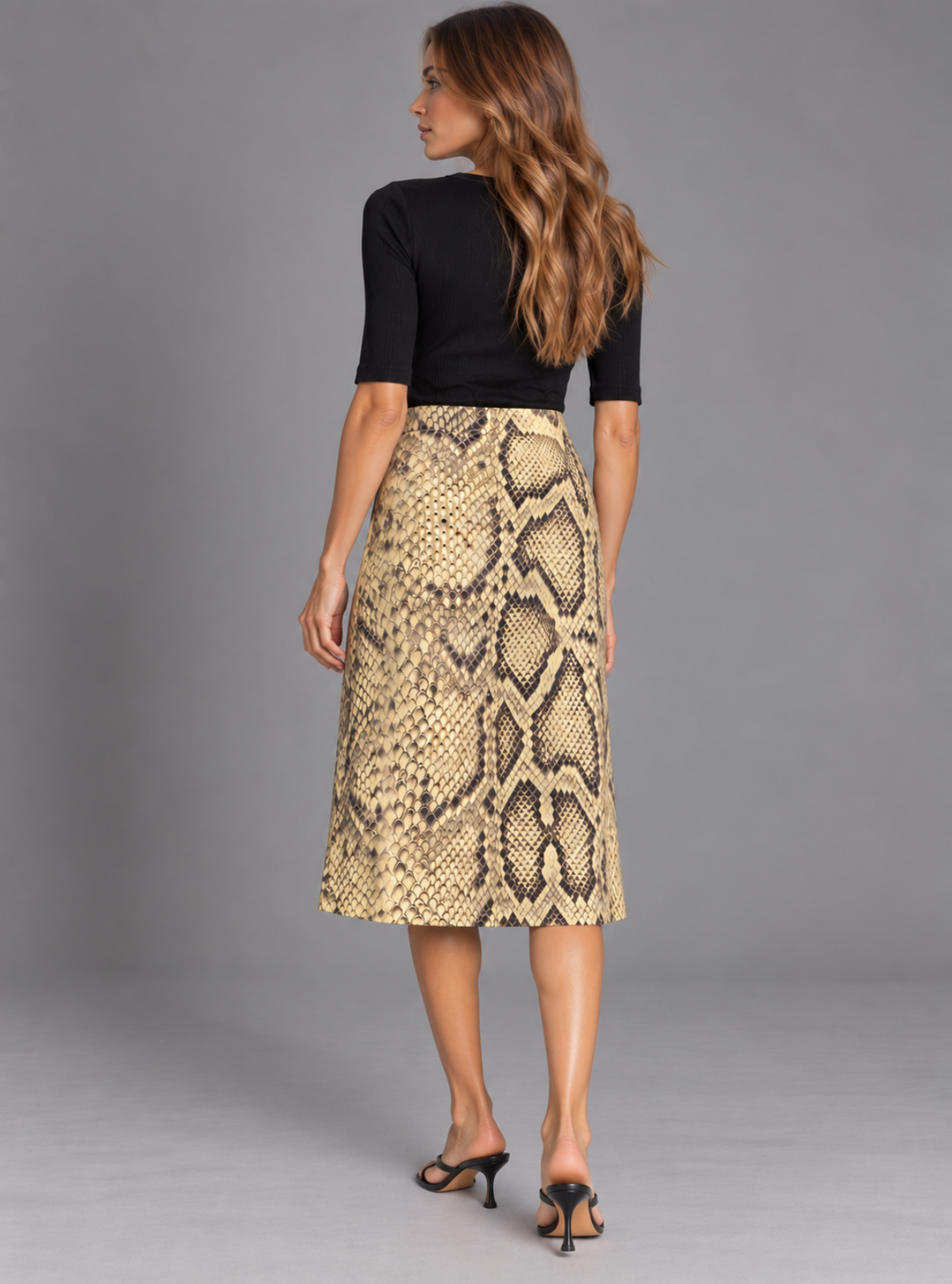A woman shown from the back wearing a black short-sleeve top and a beige and brown snakeskin print midi skirt, paired with black heeled sandals, against a solid gray studio background.